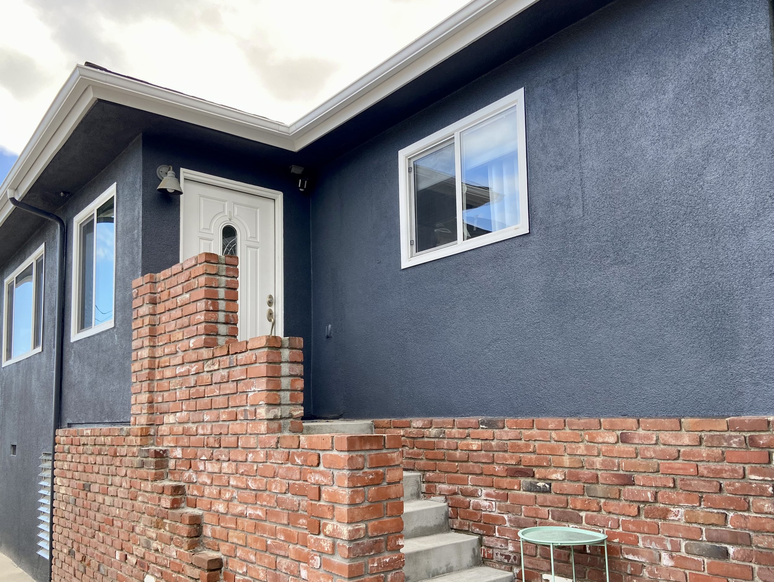 Dark blue stucco home exterior with white-trimmed windows, a decorative front door, concrete steps, and a red brick entry wall.
