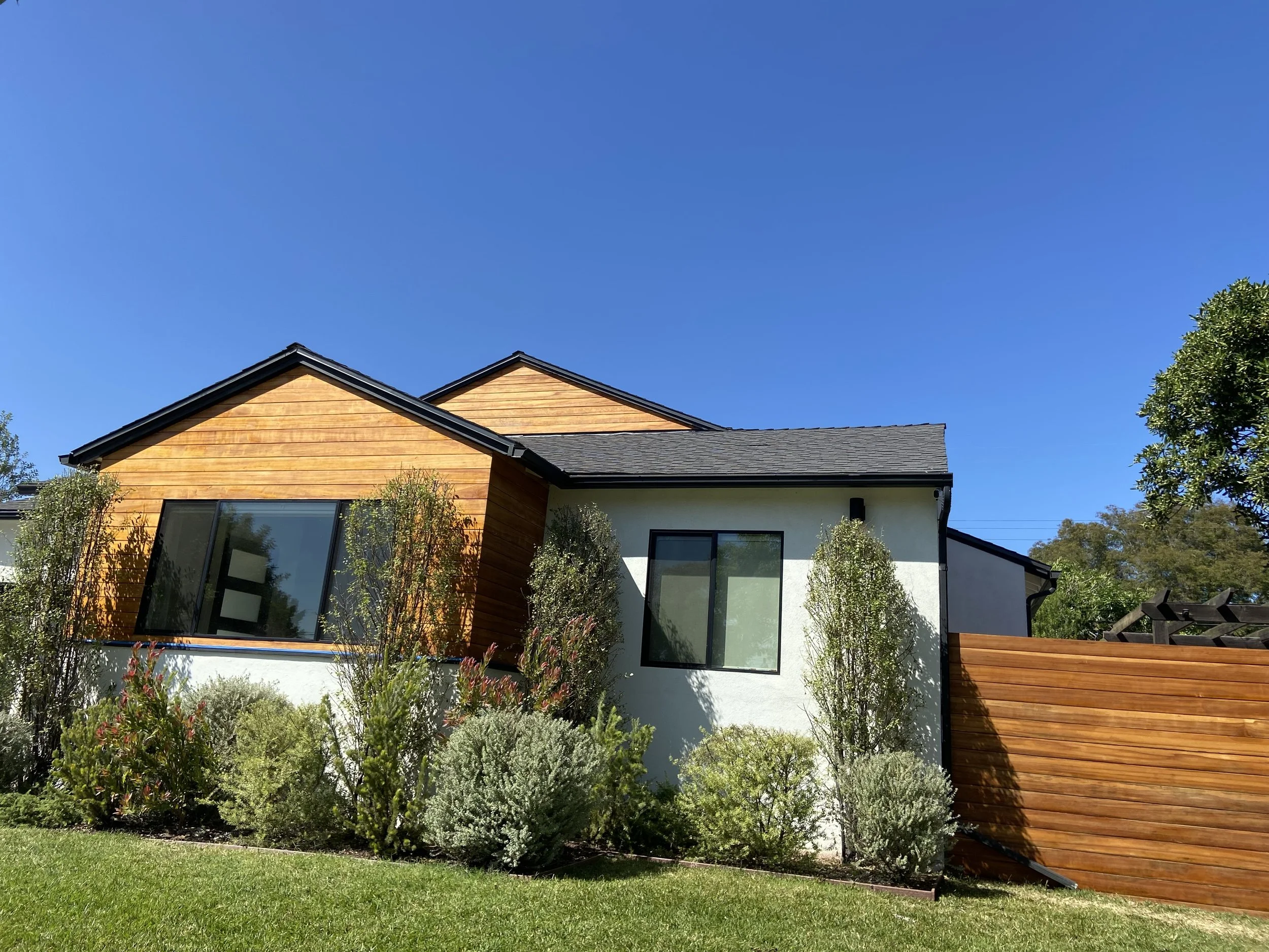 Modern single-story home with wood siding, white stucco walls, black-framed windows, landscaped yard, and wooden privacy fence under a clear blue sky