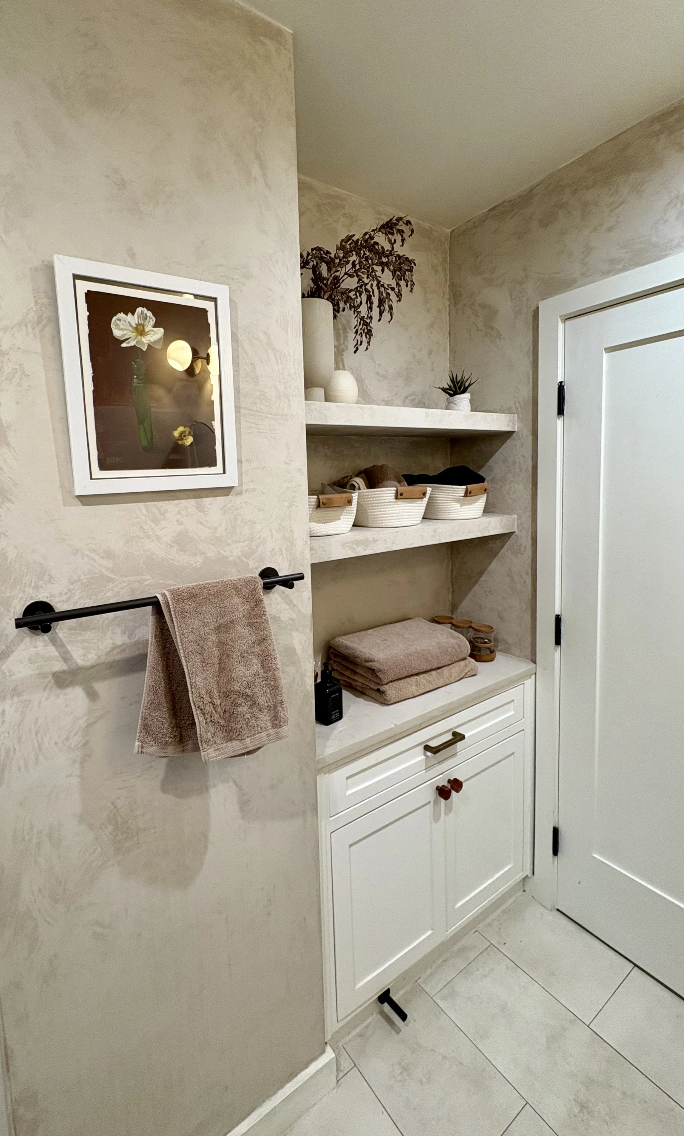 Modern beige bathroom with textured walls, white built-in shelving, woven baskets, folded towels, potted plants, framed floral artwork, and black towel bars.