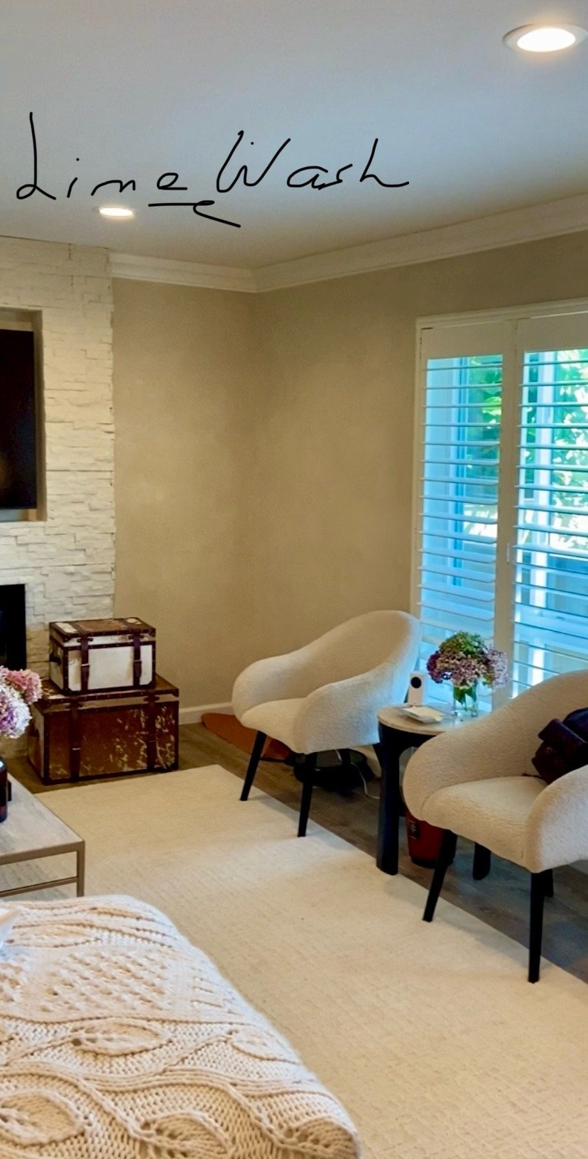 Bright modern living room with cream boucle chairs, a woven throw, floral arrangement, storage trunks, large windows, and handwritten “Lime Wash” text on the ceiling.