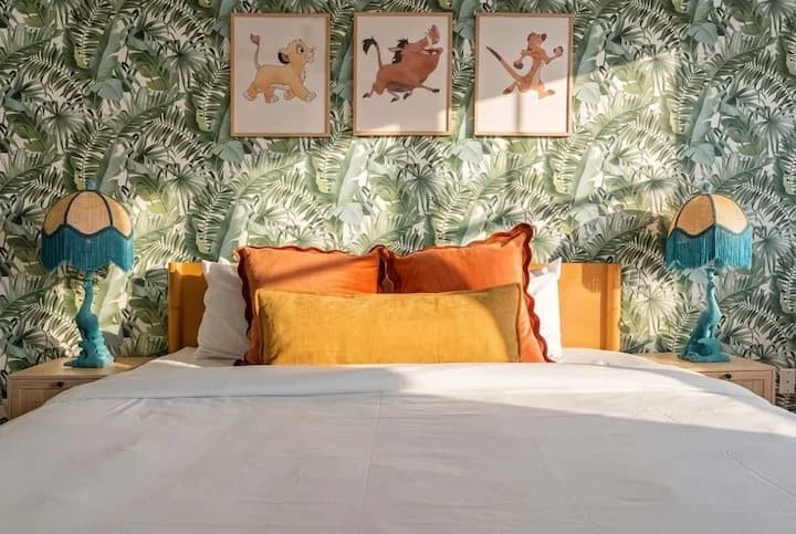 Colorful tropical bedroom with patterned wallpaper, orange and yellow pillows, blue bedside lamps, and framed animal artwork above the bed