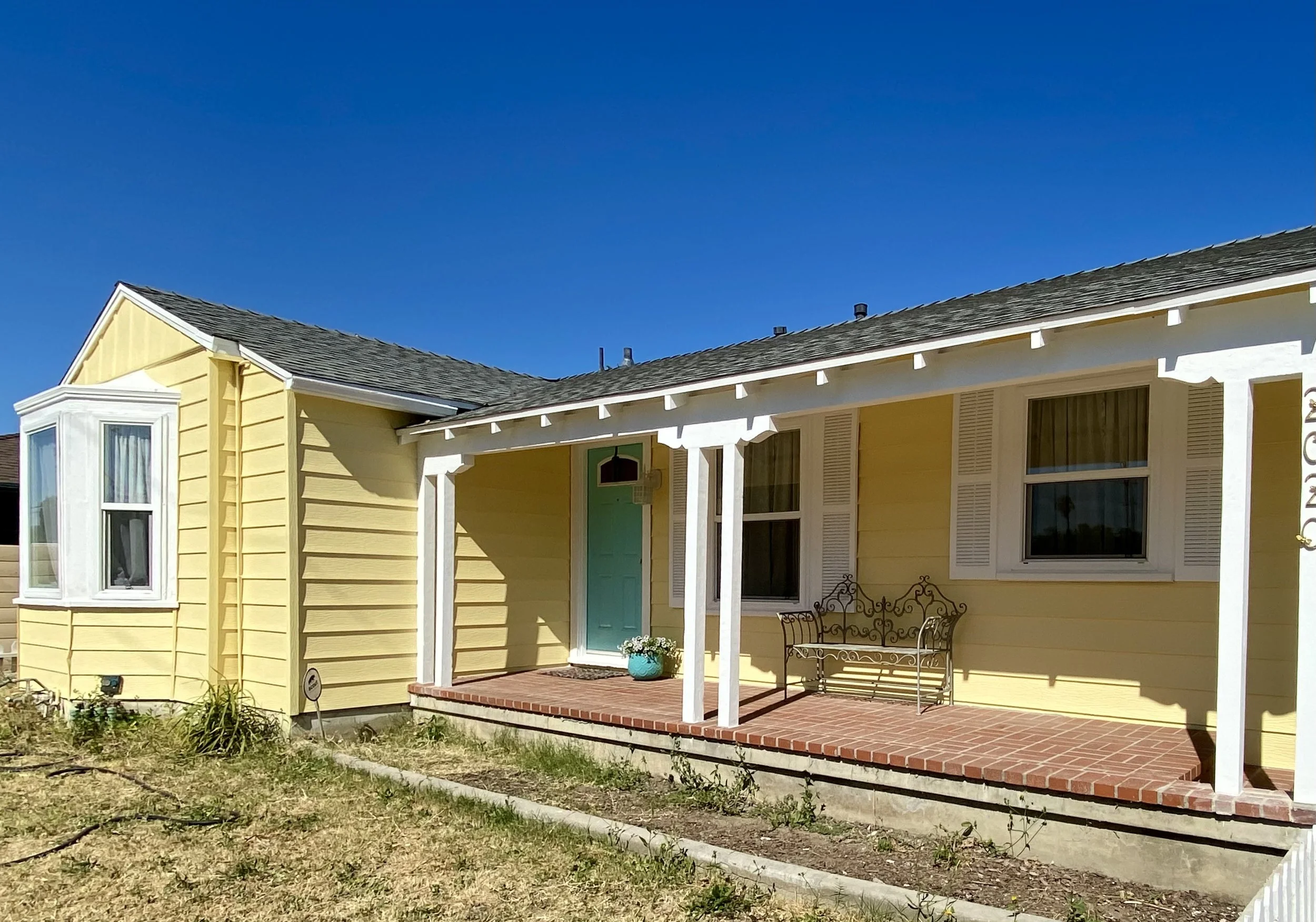 Yellow single-story home with white trim, turquoise front door, covered brick porch, and outdoor bench under a clear blue sky