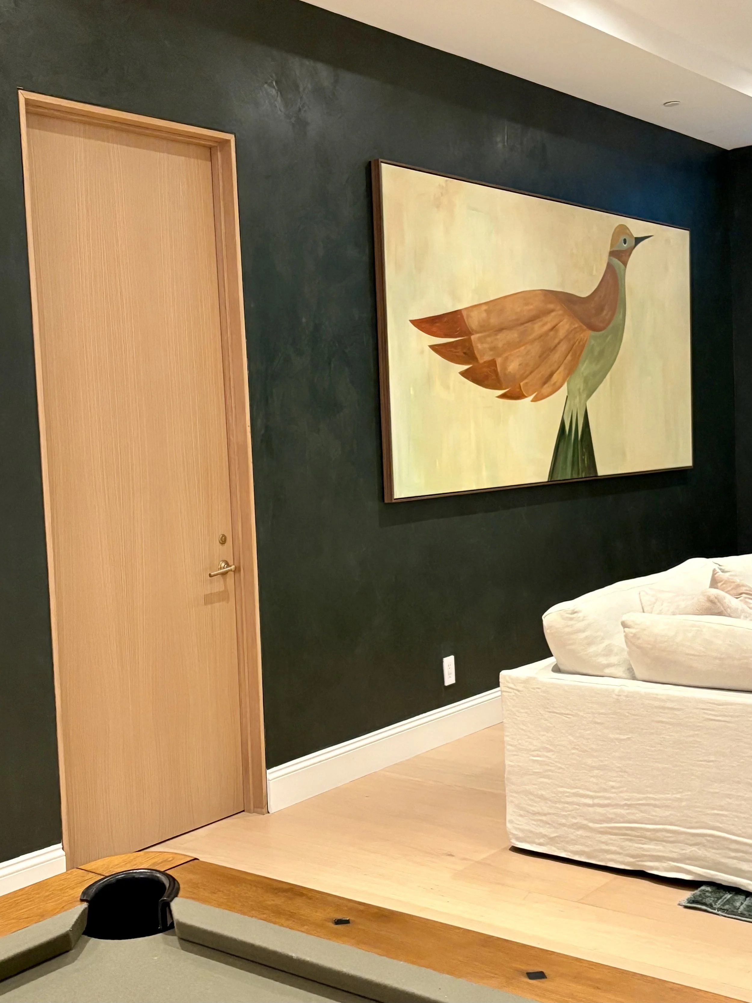Dark green textured accent wall with natural wood door, framed artwork, and cream sofa in a modern interior.