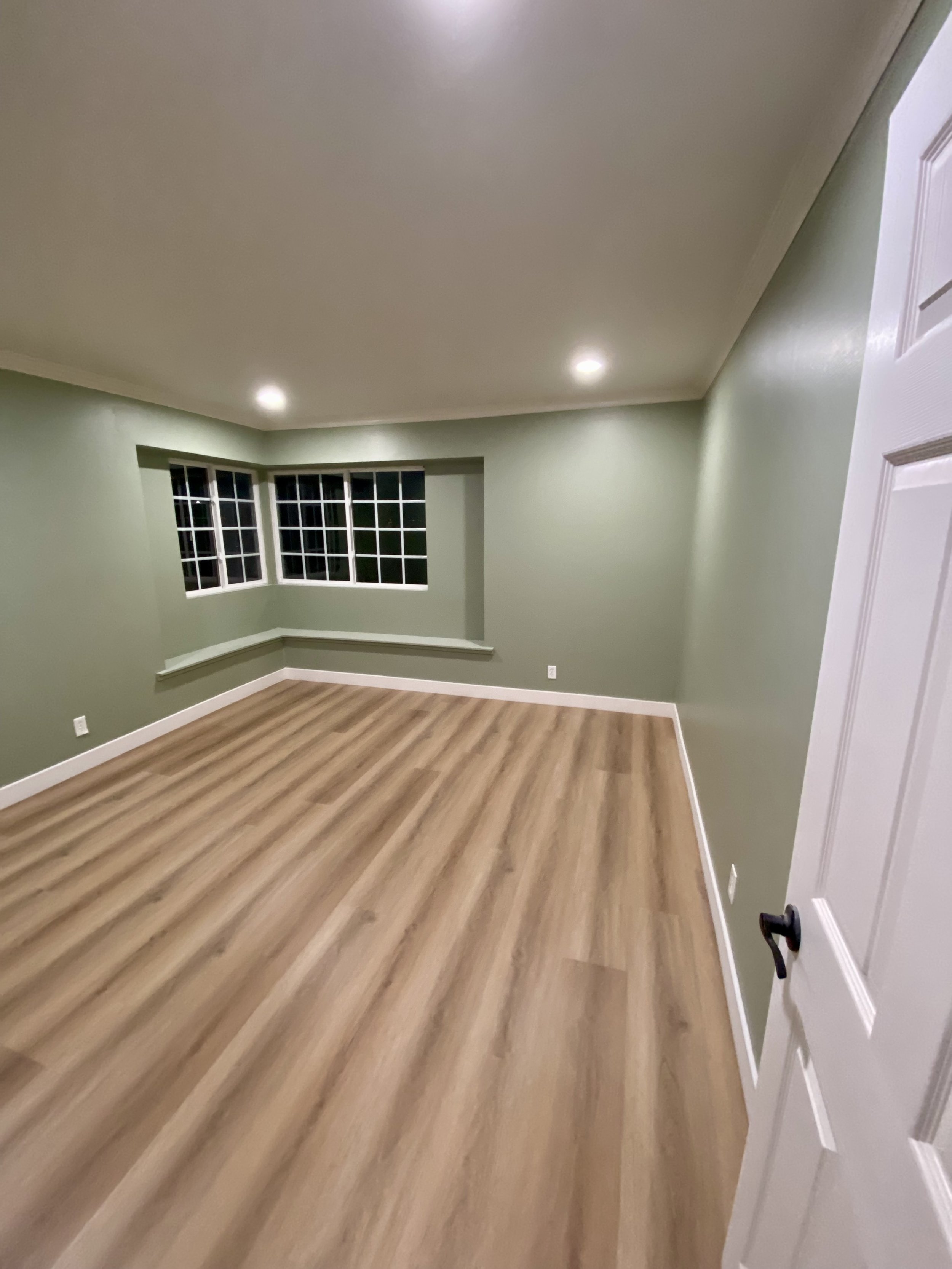 Empty green-painted room with wood-look flooring, recessed ceiling lights, white trim, and bay windows.