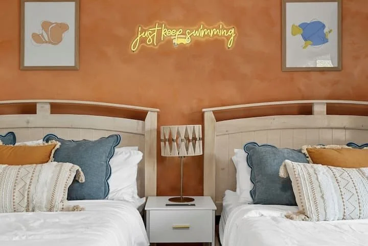 Symmetrical bedroom with twin beds, blue and white pillows, orange accents, a bedside table, and a glowing “just keep swimming” wall sign
