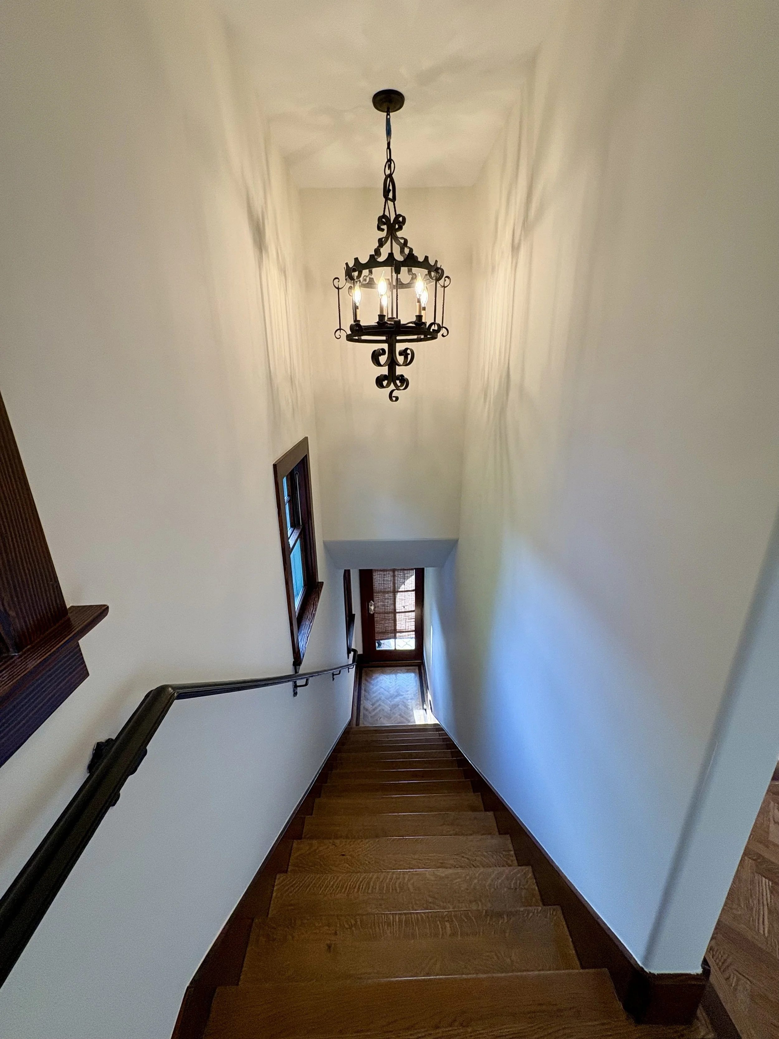 Ornate wrought-iron chandelier hanging above a wooden staircase in a bright hallway
