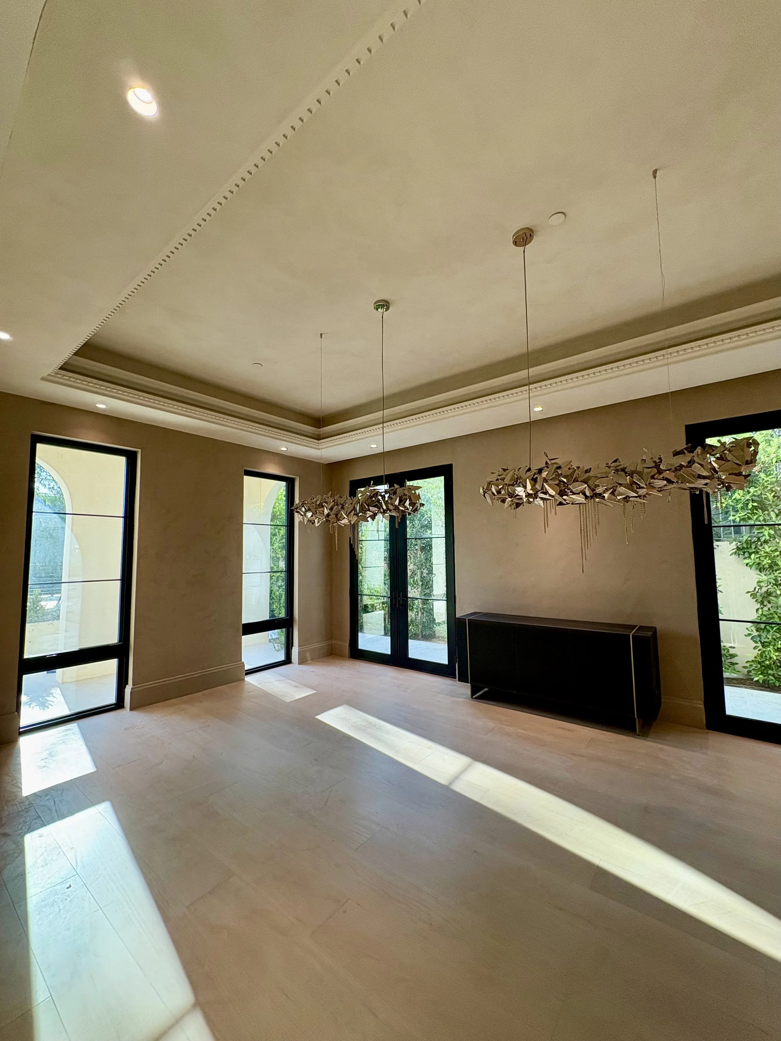 Warm taupe textured walls in a dining room with black-framed windows, sculptural chandeliers, and decorative ceiling molding.