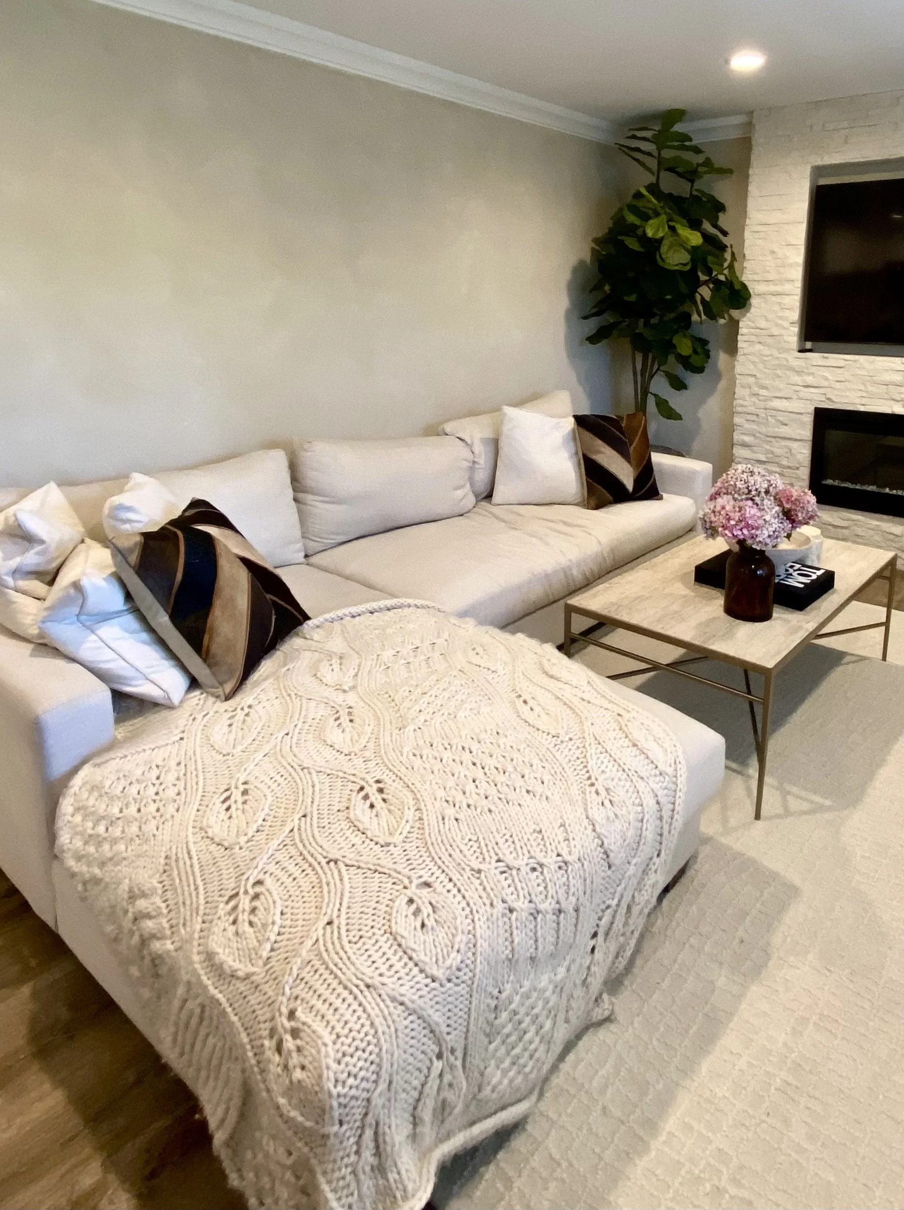 Modern living room with a white sectional sofa, textured knit ottoman, coffee table, potted plant, and stone fireplace