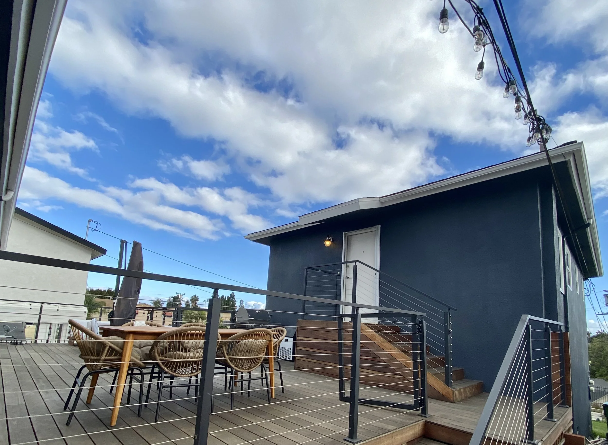Modern rooftop deck with outdoor dining furniture, grill, cable railing, string lights, and a dark blue house under a partly cloudy sky