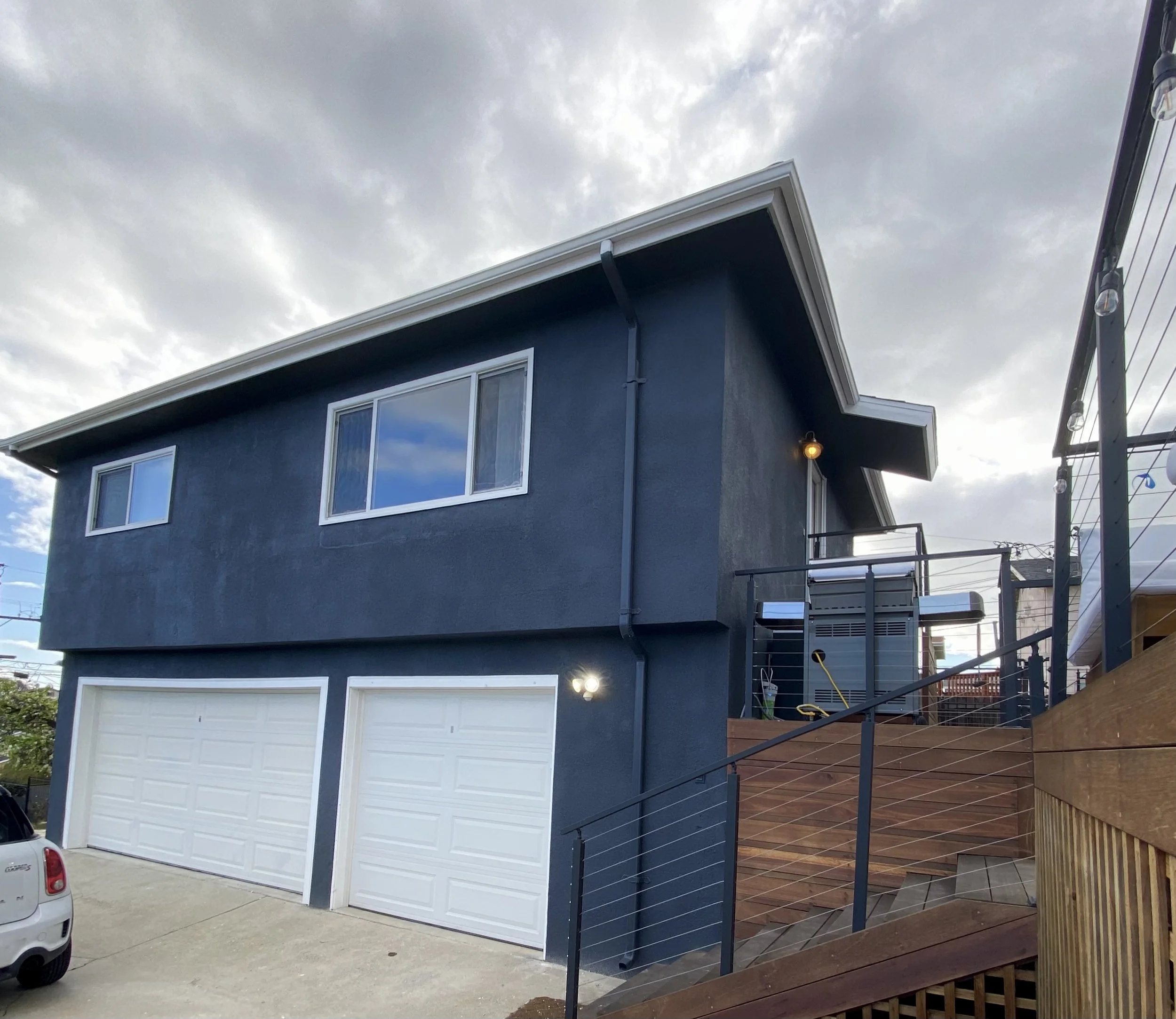 Dark blue two-story house with white-trimmed windows, two garage doors, and a wooden deck under a cloudy sky