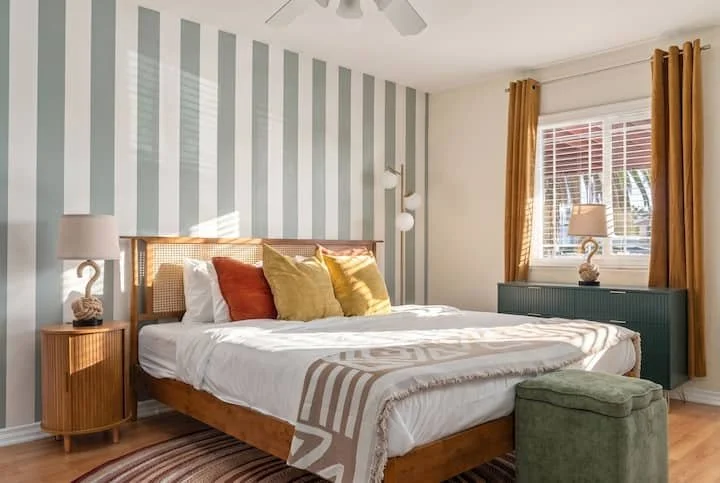 Bright modern bedroom with a wooden bed, striped accent wall, colorful pillows, mustard curtains, and green ottoman