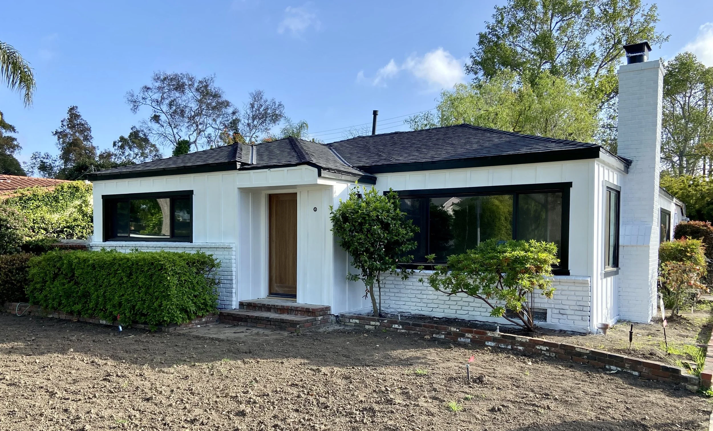 White mid-century modern house with black-framed windows, landscaped shrubs, brick planters, and a tall chimney