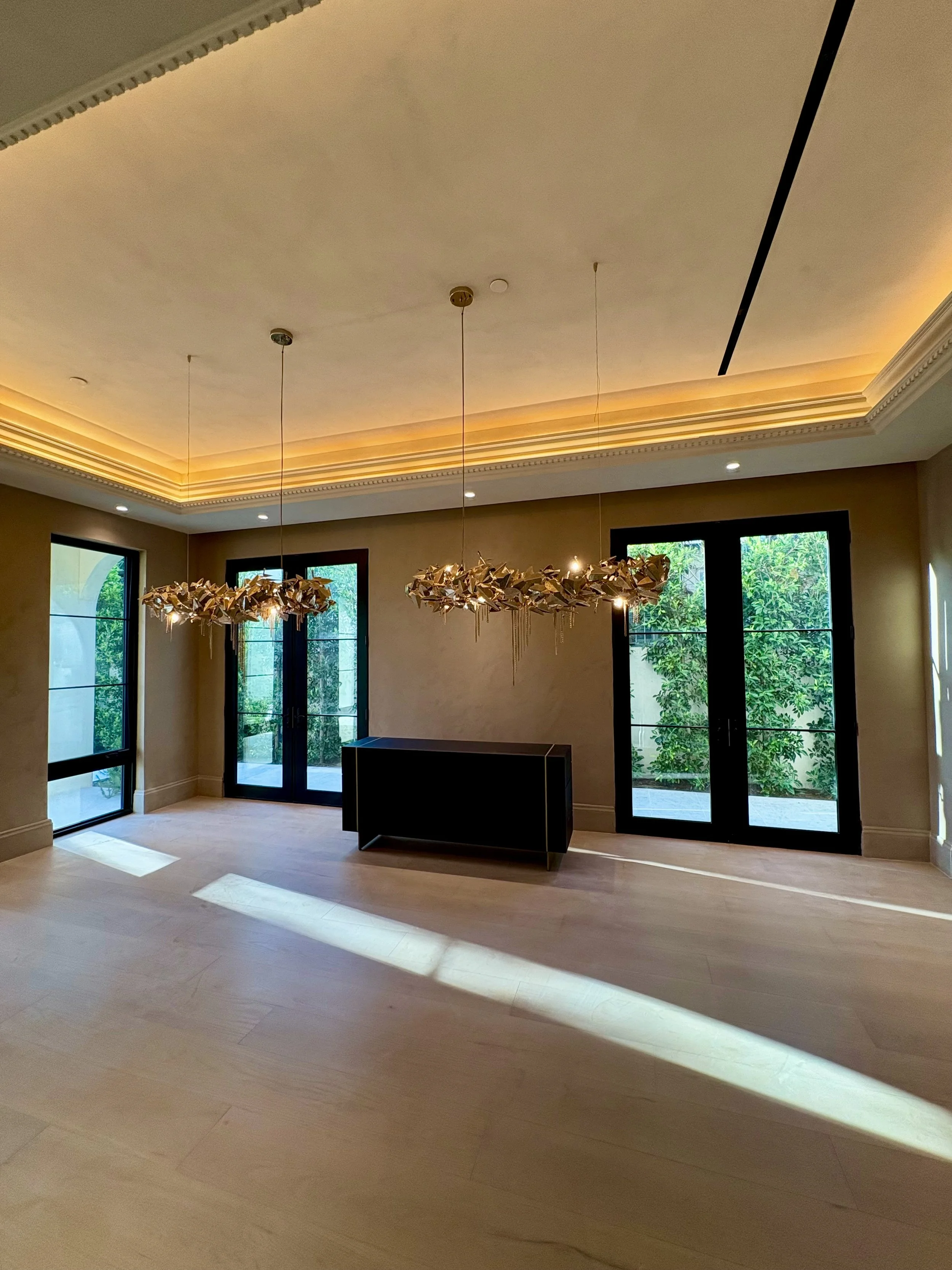 Elegant empty interior with ornate crown molding, illuminated tray ceiling, sculptural leaf chandeliers, black-framed windows, and a black modern fireplace.
