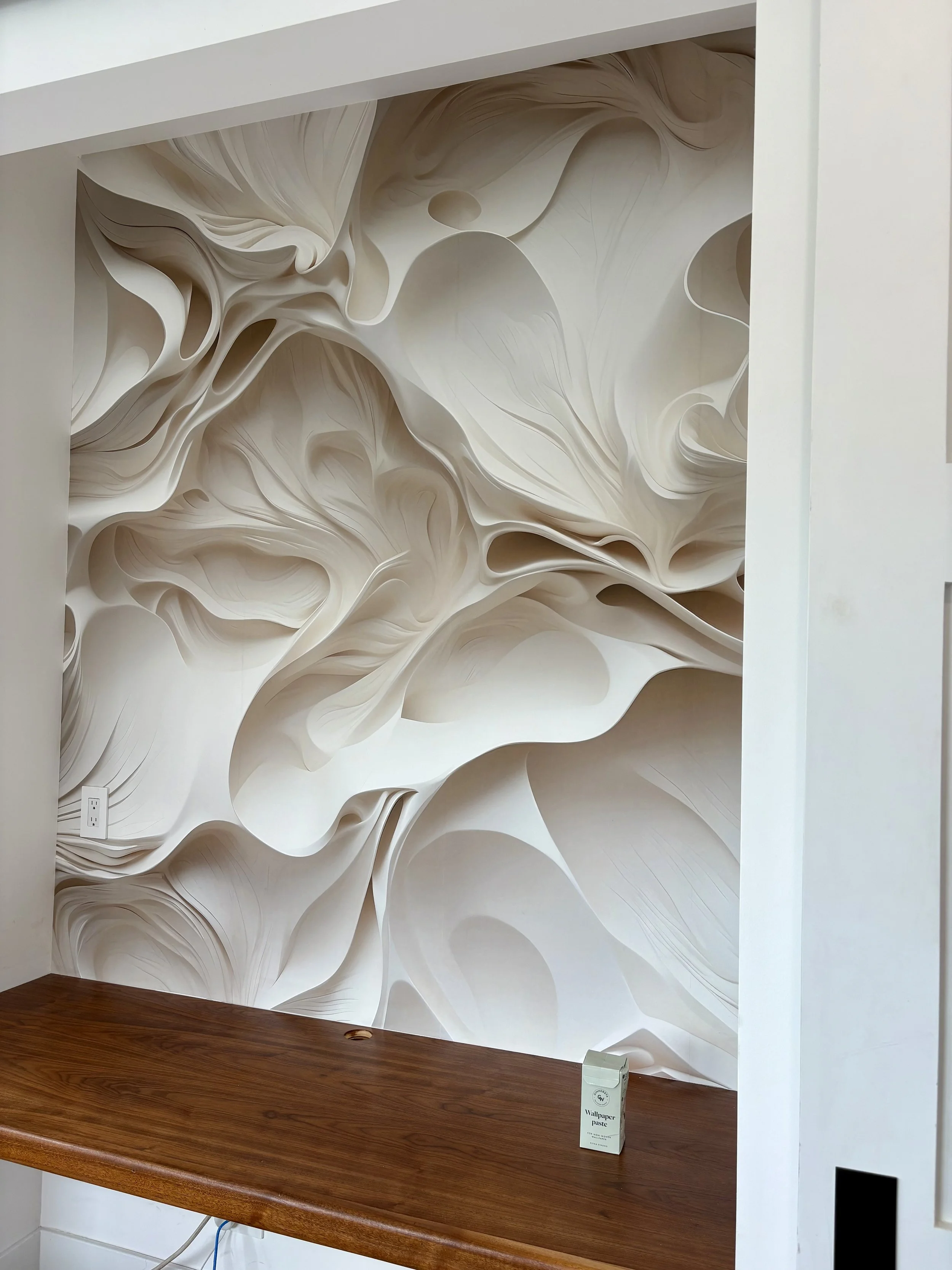 Close-up of abstract cream-and-white sculptural wallpaper with flowing organic patterns above a wooden mantel, with wallpaper paste on the shelf