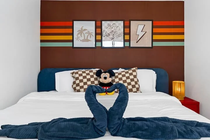 Mickey Mouse plush toy sitting on a neatly made bed with blue blankets, striped wall art, and bedside lamp