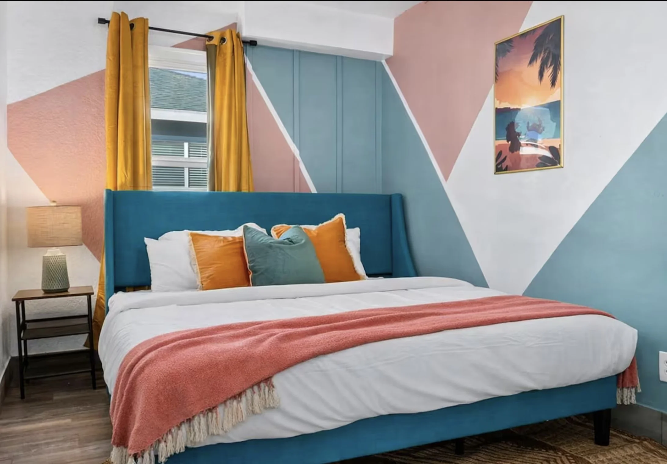 Colorful modern bedroom with a teal upholstered bed, white bedding, coral throw blanket, orange and teal pillows, mustard curtains, geometric accent wall, and tropical artwork.