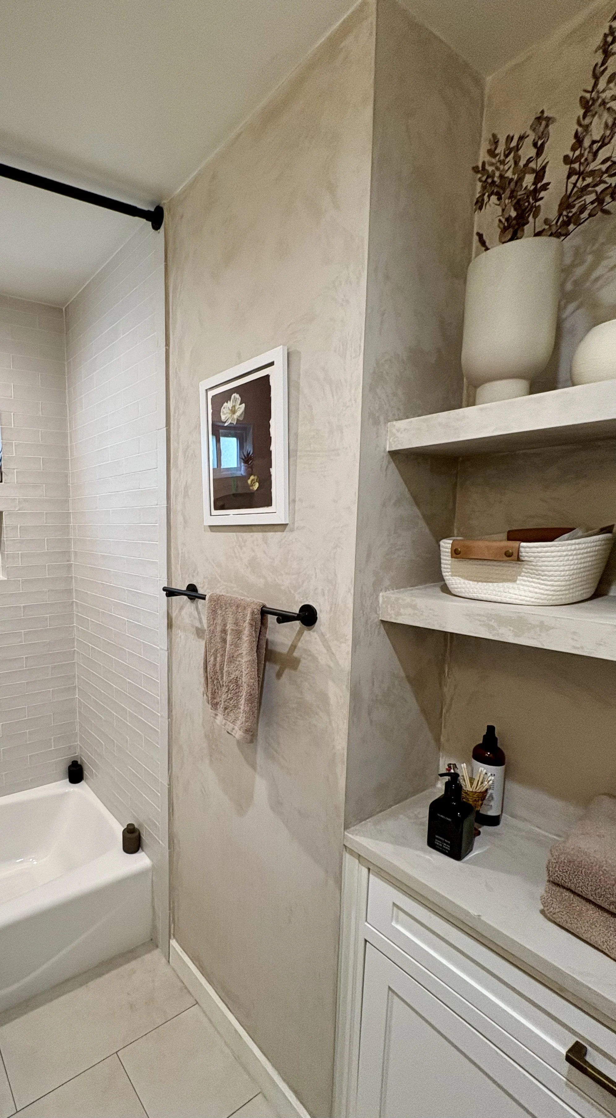 Bright modern bathroom with a bathtub, white subway tile, built-in shelves, toiletries, towels, and framed floral artwork
