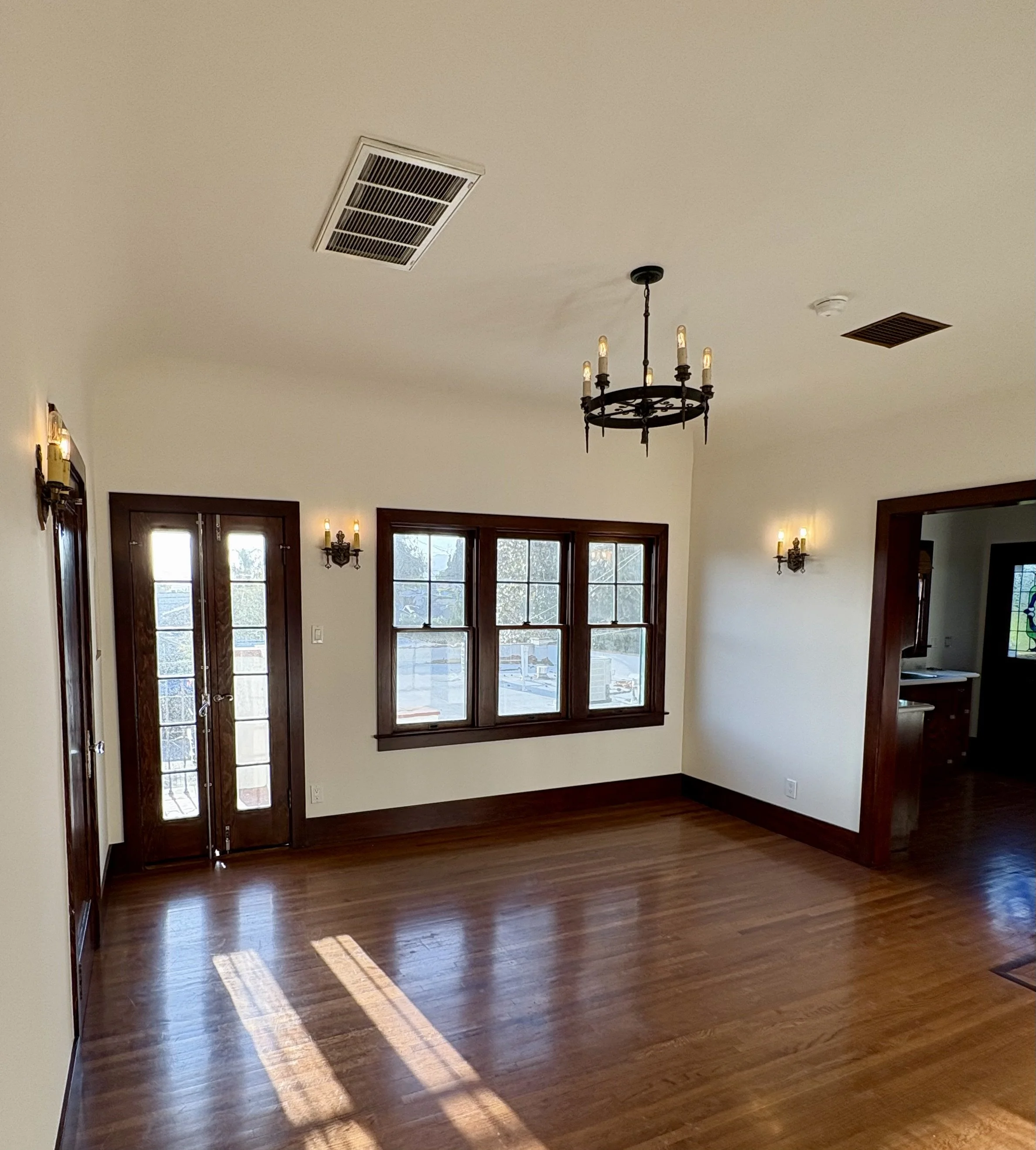 Empty historic dining room with polished hardwood floors, dark wood trim, large windows, French doors, wall sconces, and a candle-style chandelier