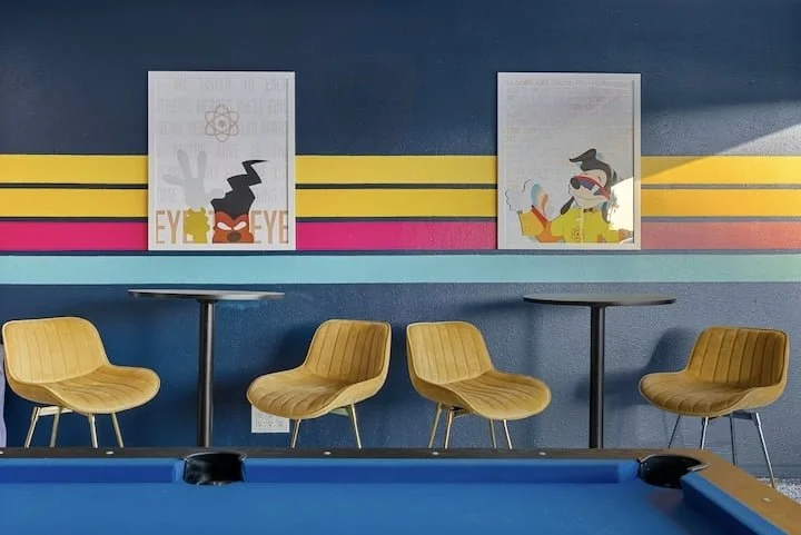Colorful retro game room with a blue pool table, four mustard-yellow chairs, round tables, and framed artwork on a striped wall