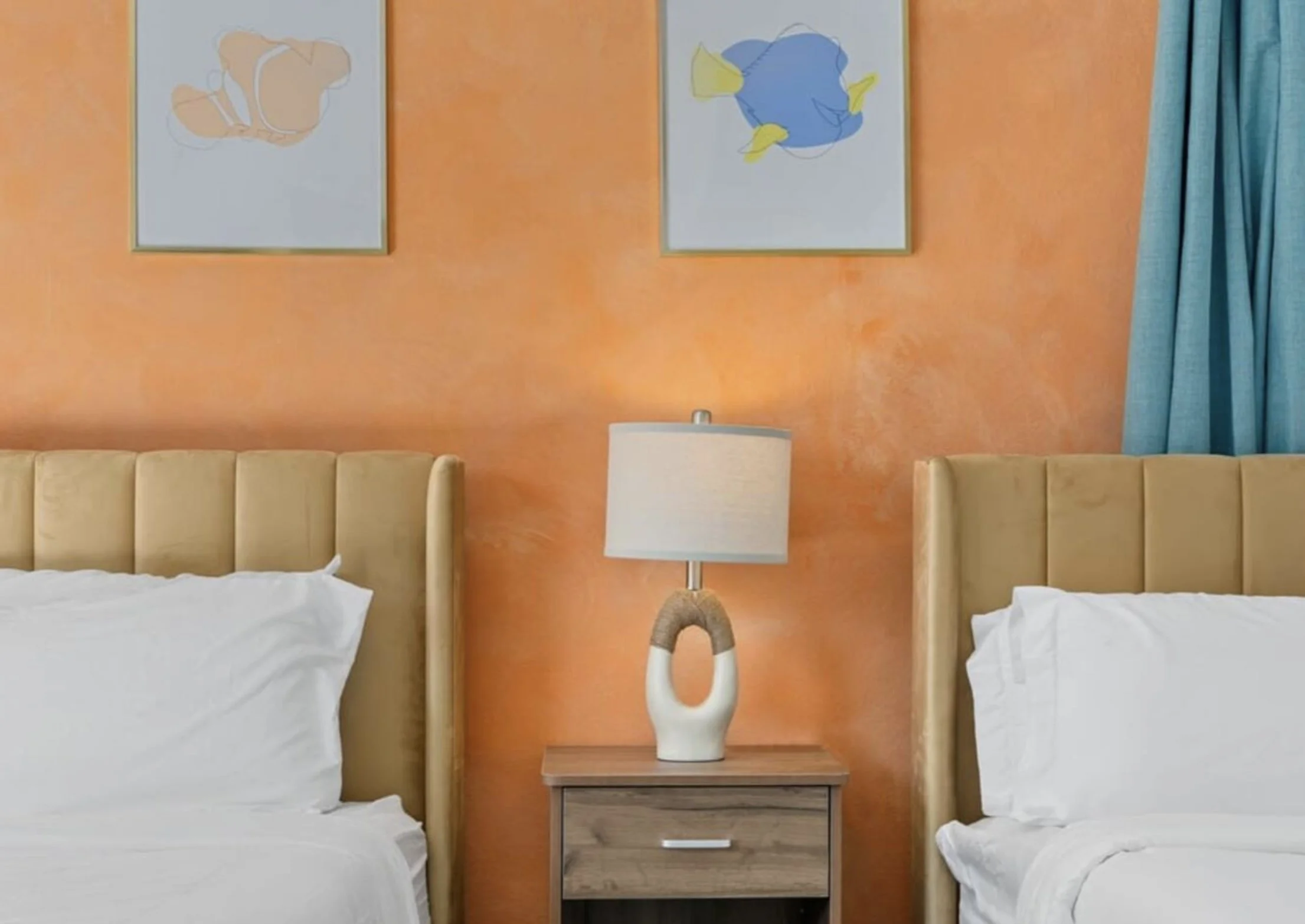 Hotel room with two twin beds, orange accent wall, bedside table, lamp, framed artwork, and blue curtains