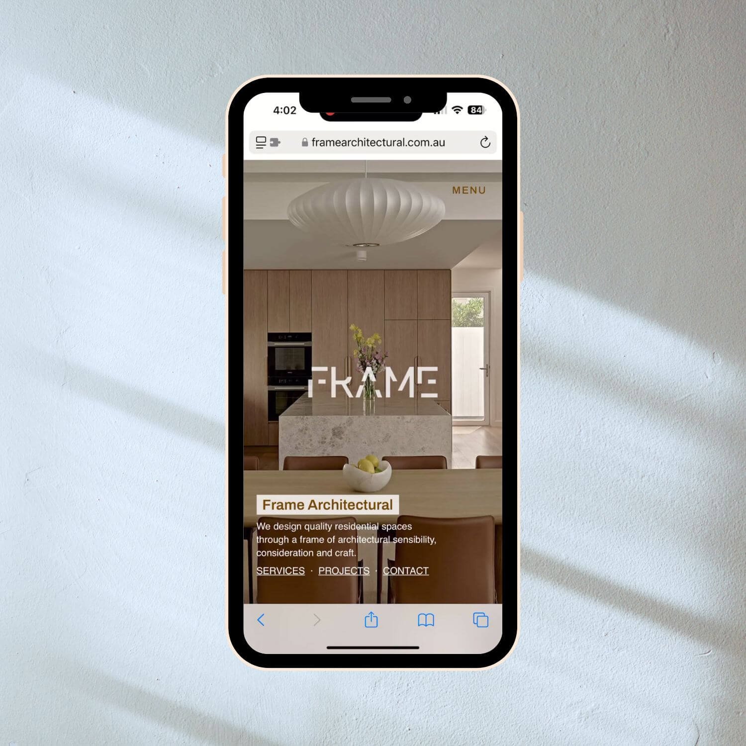 Smartphone displaying the Frame Architectural website homepage, featuring a modern kitchen with wood cabinetry, a central island, and a large white pendant light. The page includes navigation links for services, projects, and contact information.