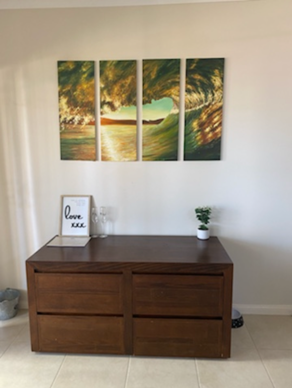 A wooden sideboard with decorative items including a framed 'love' sign, and a small plant, against a white wall. Above, a multi-panel painting of a large ocean wave at sunset.