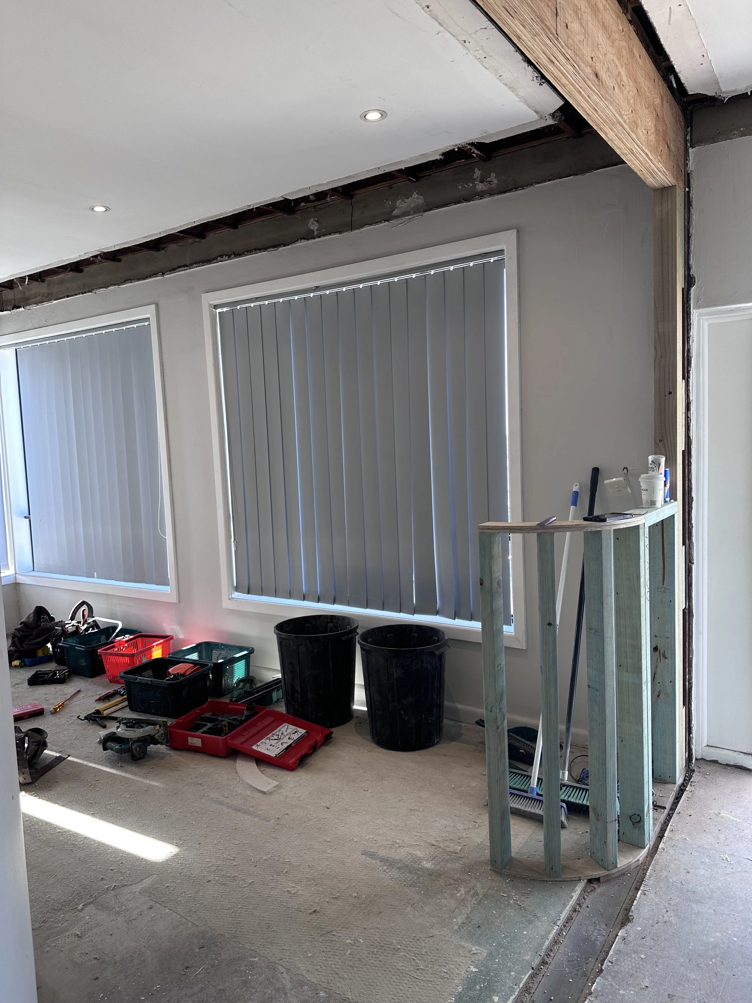 Room under renovation with tools and materials, window with vertical blinds, partial wall framing, exposed ceiling structure, and construction supplies on the floor.