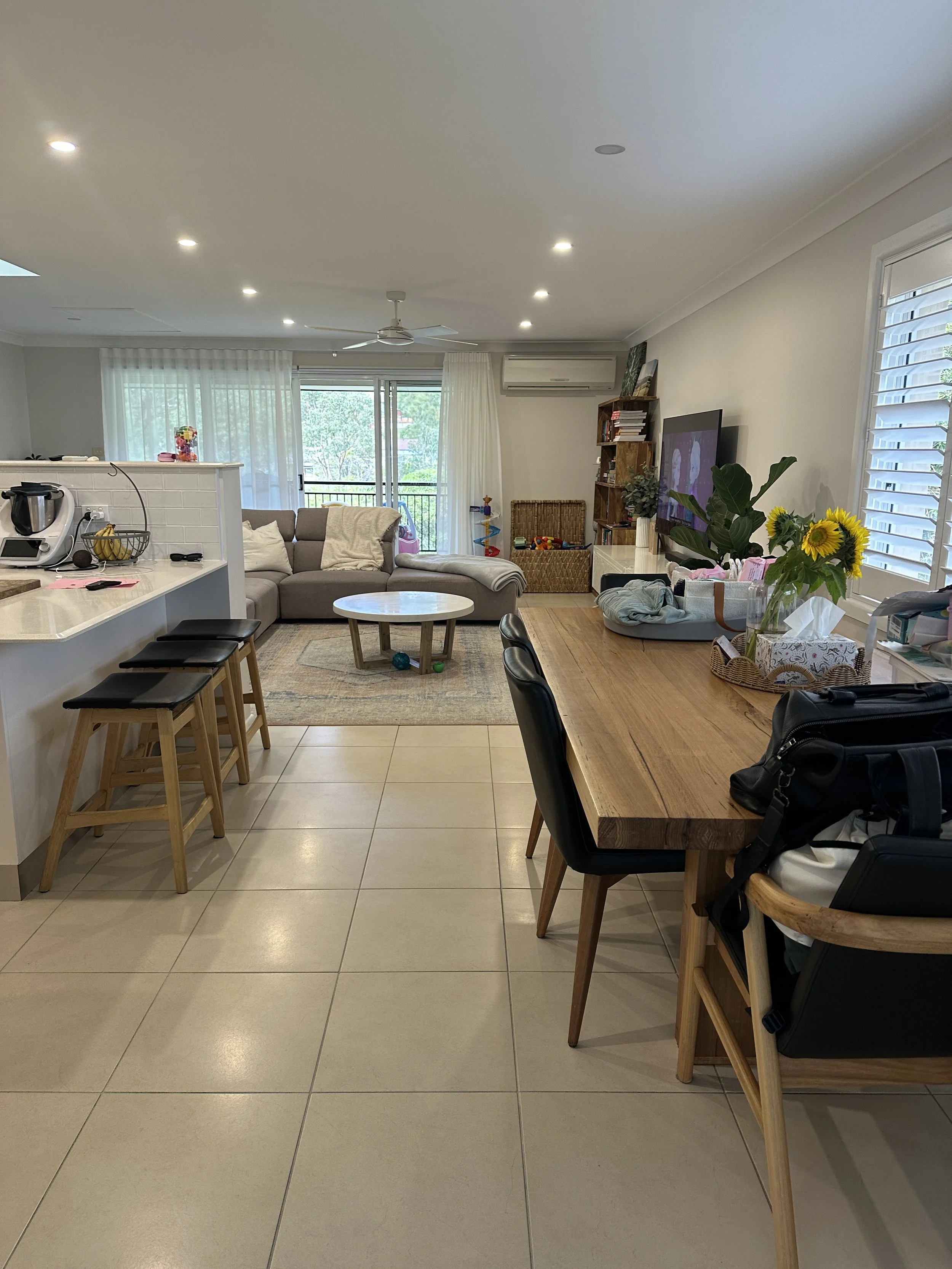 Open-concept living room and kitchen with beige tiled floors, wooden dining table, black chairs, and a kitchen counter with two black stools. The living area has a gray sectional sofa, a round coffee table, a TV, bookshelves, and sliding glass doors 