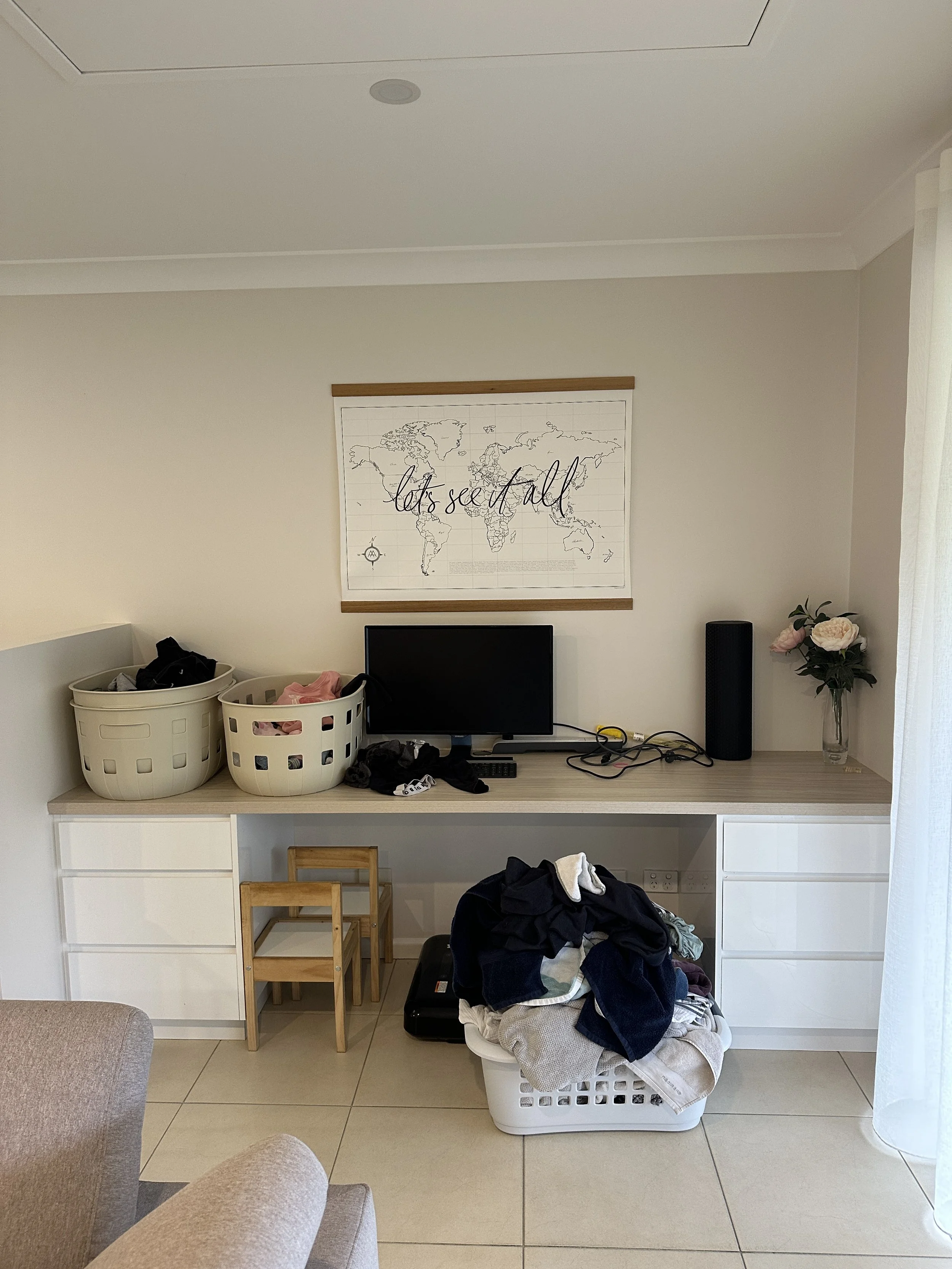 Living room or workspace with a white desk, a small monitor, laundry baskets with clothes, a basket with laundry, two small wooden chairs, a potted plant with pink flowers, and a white curtain.