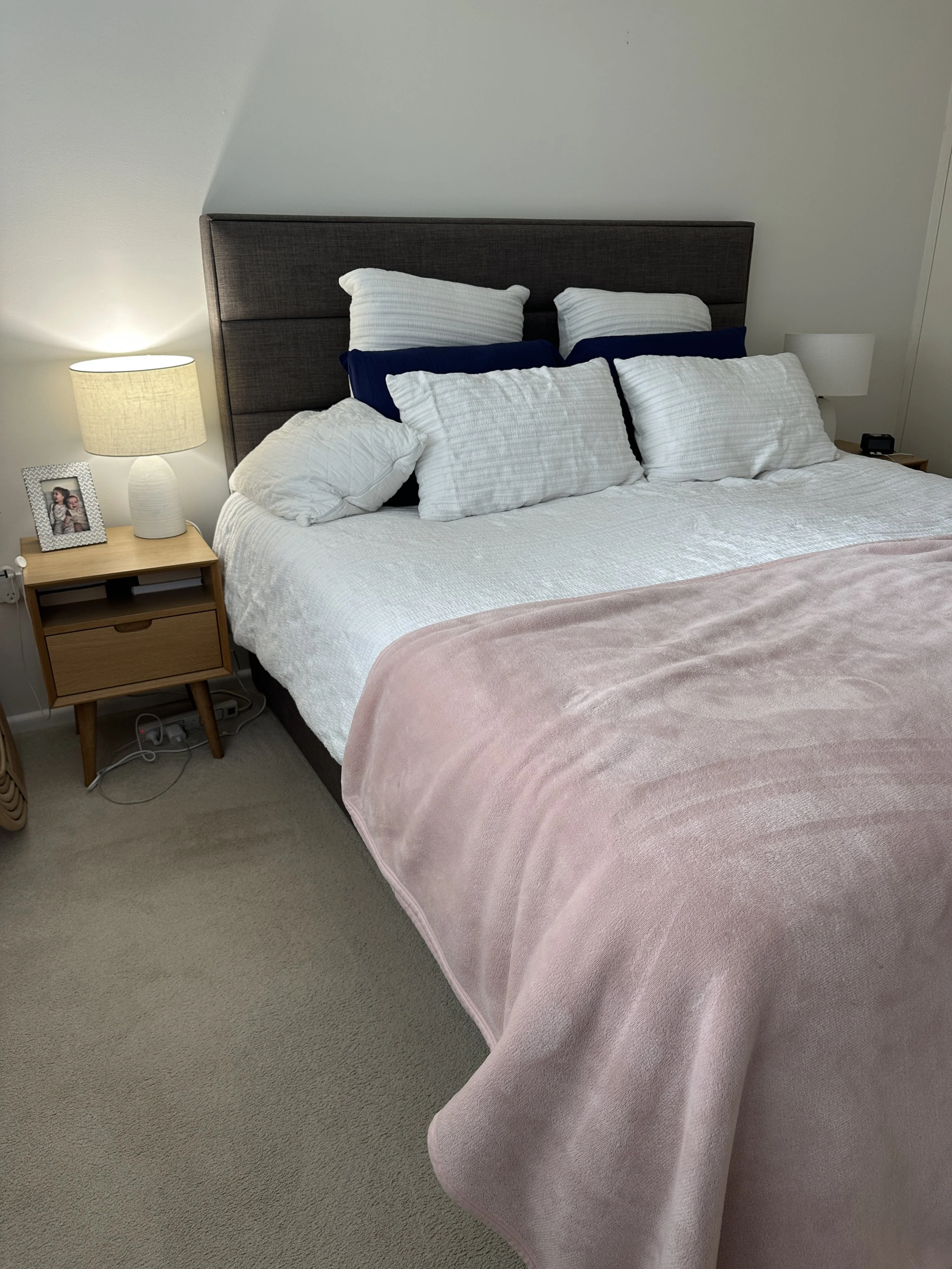 A neatly made bed with white, blue, and striped pillows, a pink blanket, and a dark upholstered headboard in a bedroom, with two bedside lamps and a picture frame on the left nightstand.