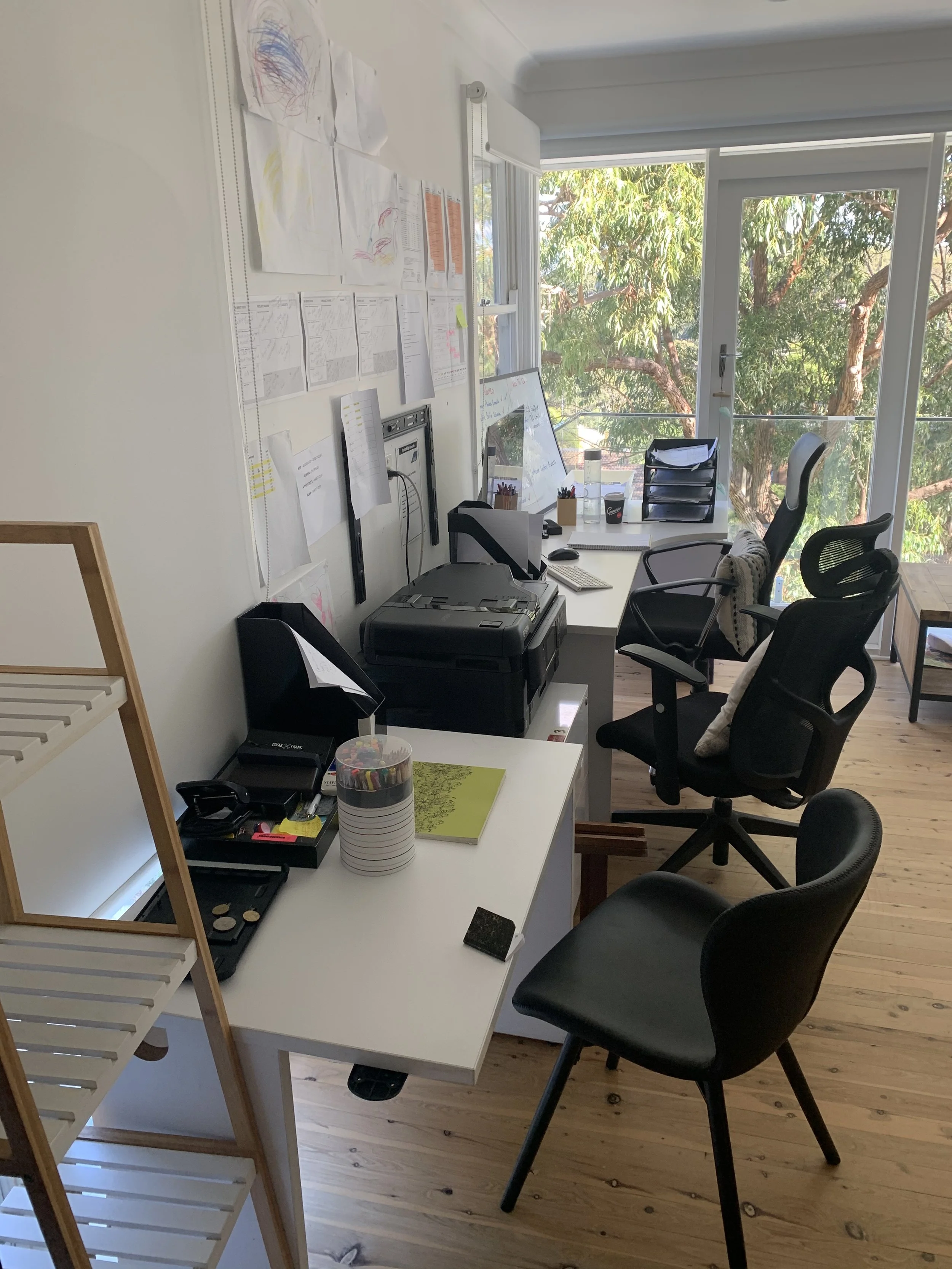 Office with two desks, chairs, a window with a view of trees, and wall with papers and drawings.