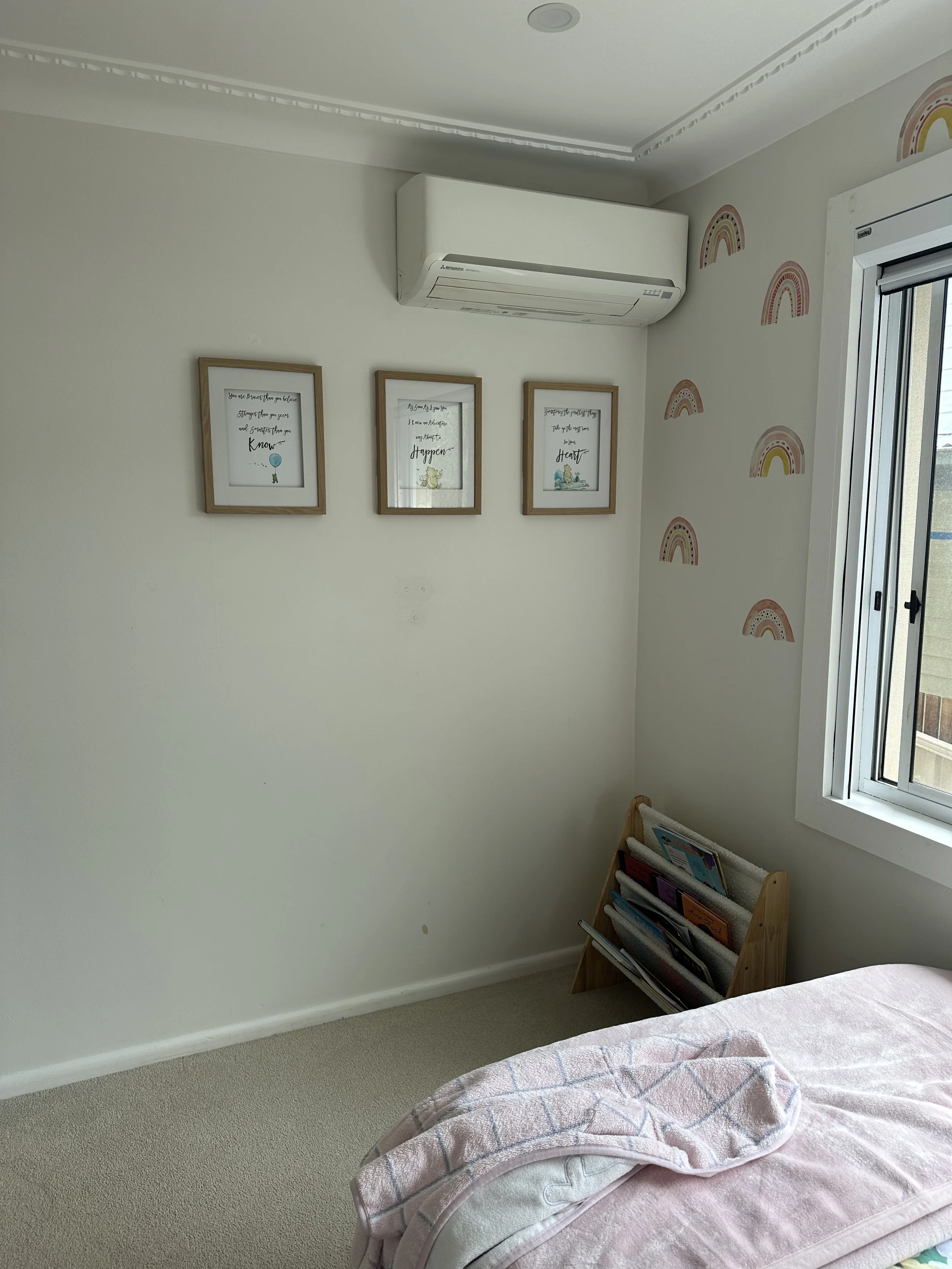 A corner of a bedroom with three framed prints on the wall, a small wooden bookshelf with magazines, and part of a bed with a pink blanket.