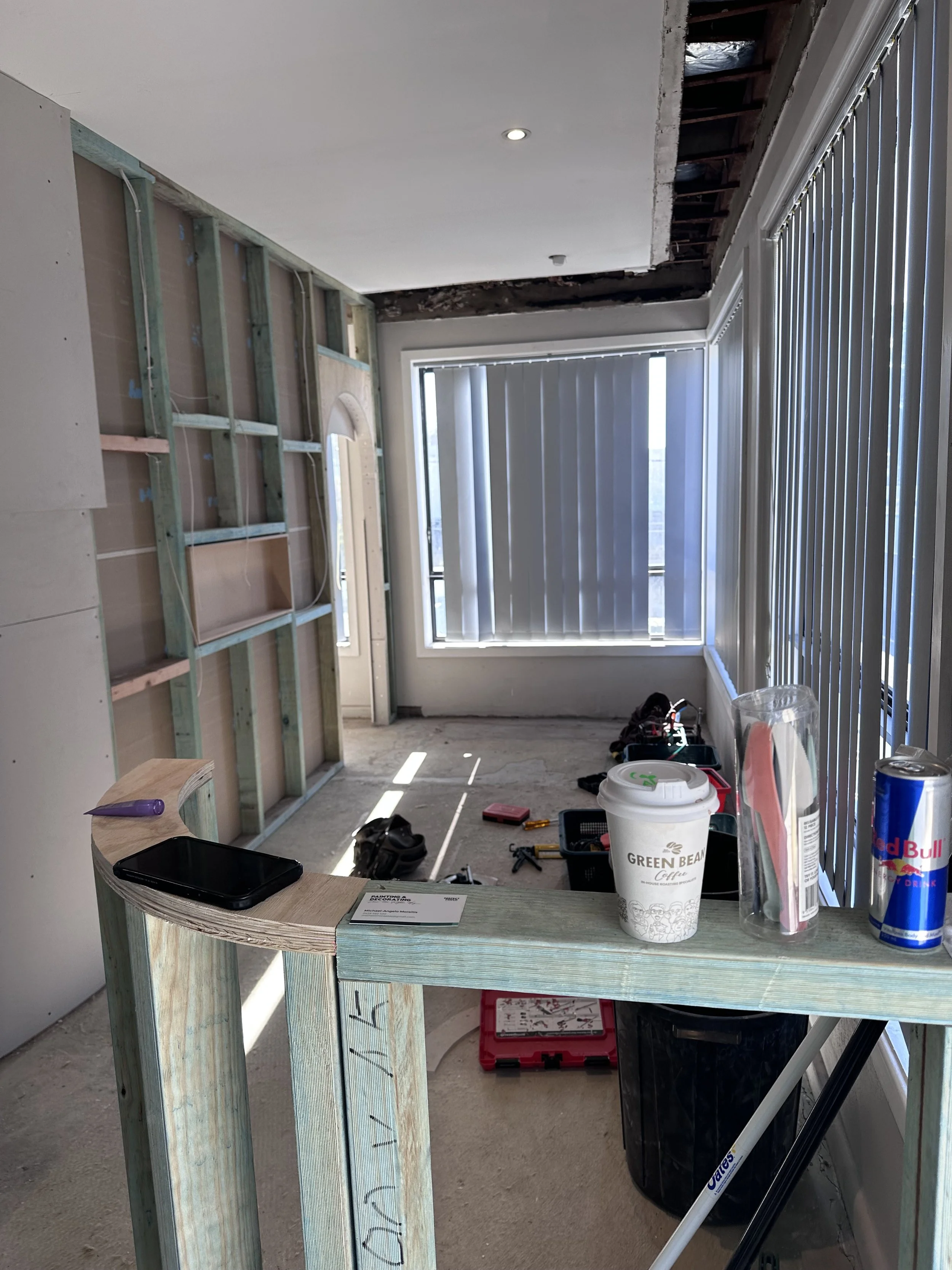 Interior of a room under renovation with exposed drywall and structural supports on one wall, large windows with vertical blinds, and a cluttered work surface with tools, a coffee cup, and miscellaneous items.