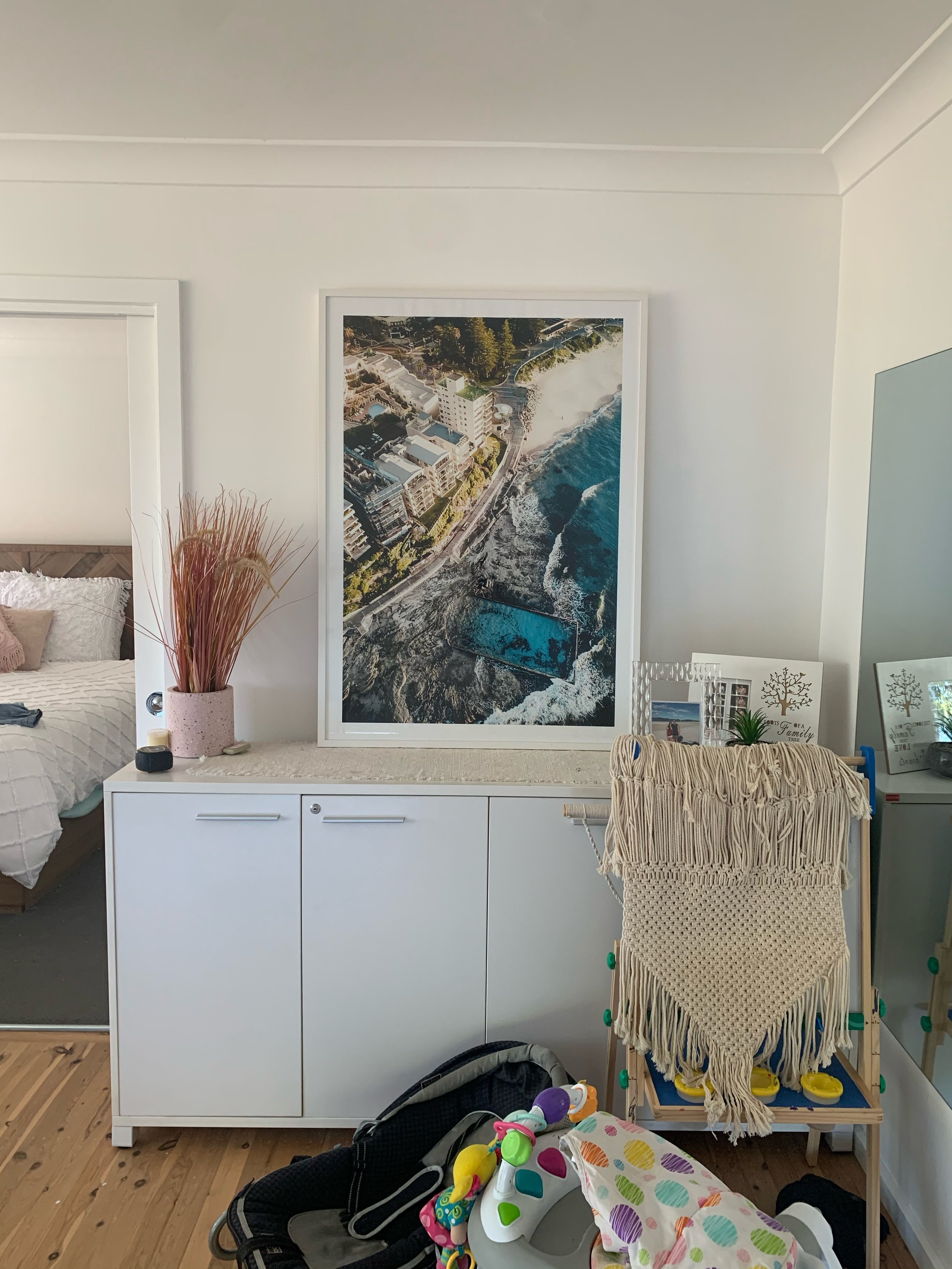 A white sideboard against a white wall with a large aerial photograph of a coastal city and a swimming pool hanging above it. To the left, part of a bed and a vase with dried pinkish plants are visible. To the right, a mirror, framed photos, a small 