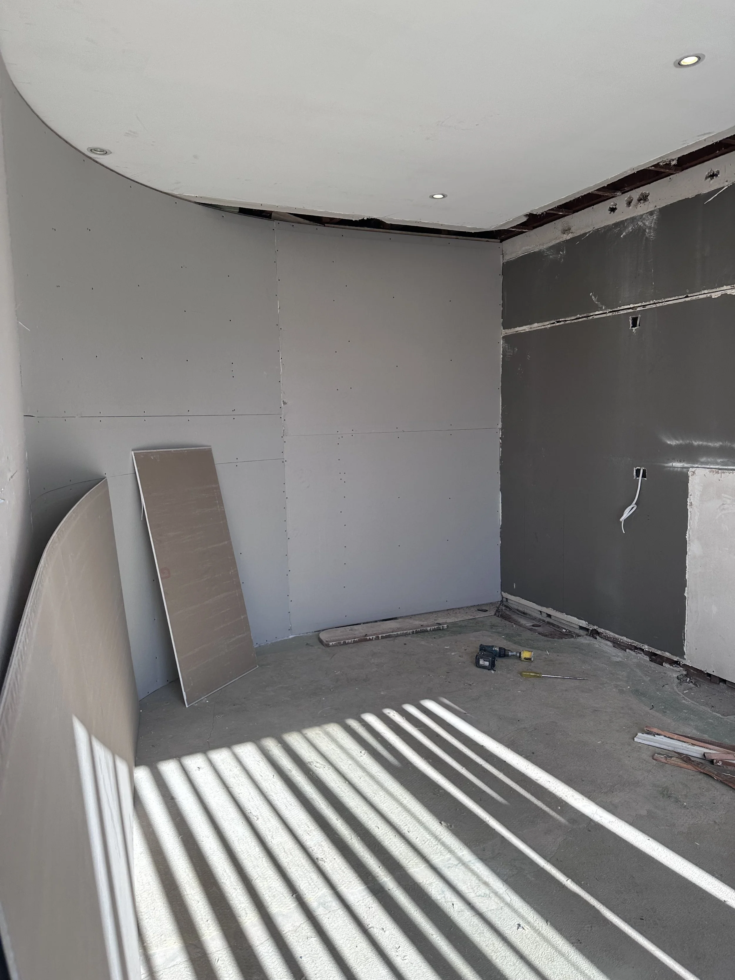 Room under construction with drywall walls, tools on the floor, and sunlight casting shadows through window blinds.
