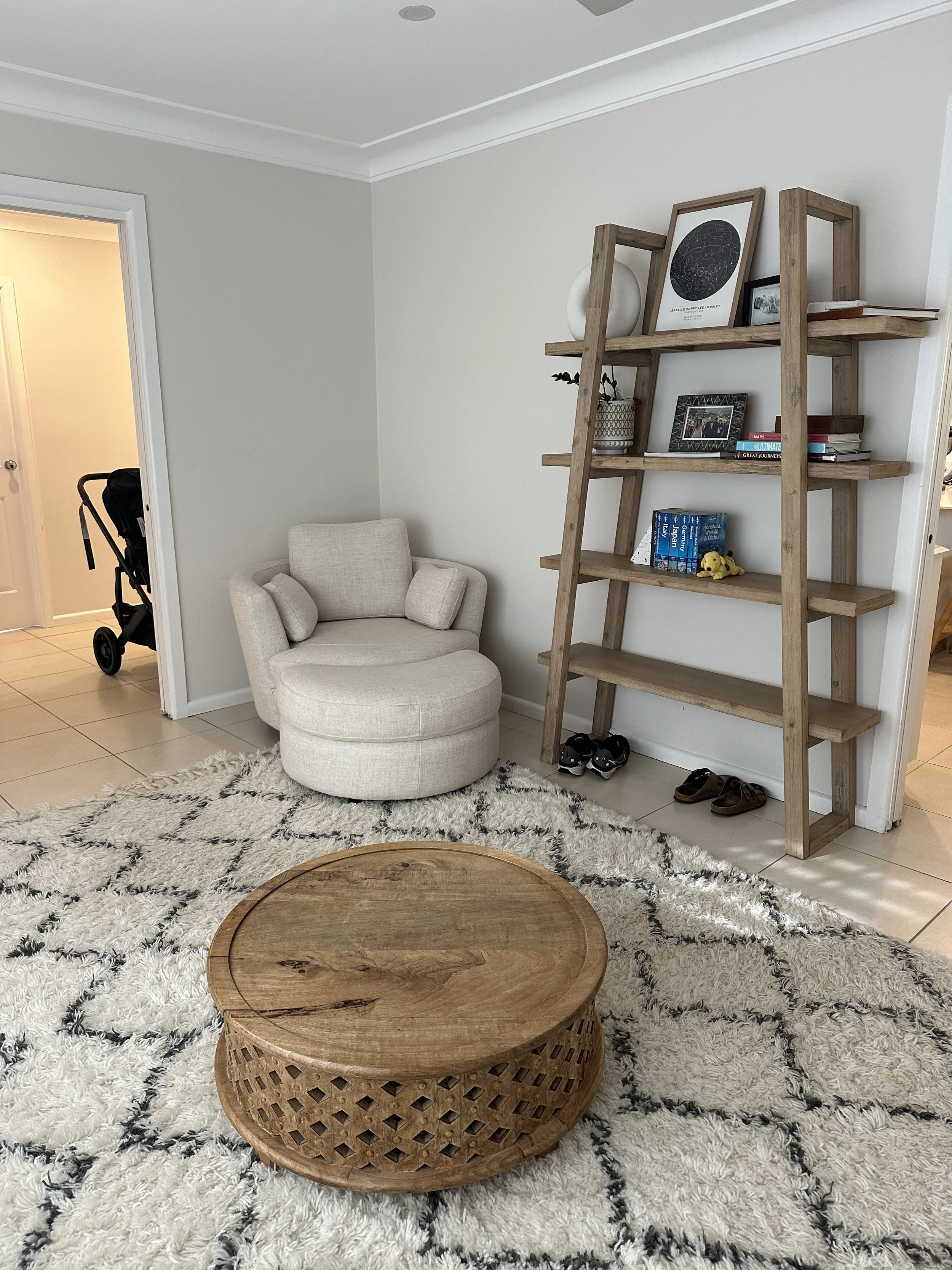 Living room corner with a round beige armchair, a wooden shelf leaning against the wall with decorative items and books, a pair of shoes under the shelf, a white shaggy rug with black diamond patterns, and a rustic wooden coffee table in front.