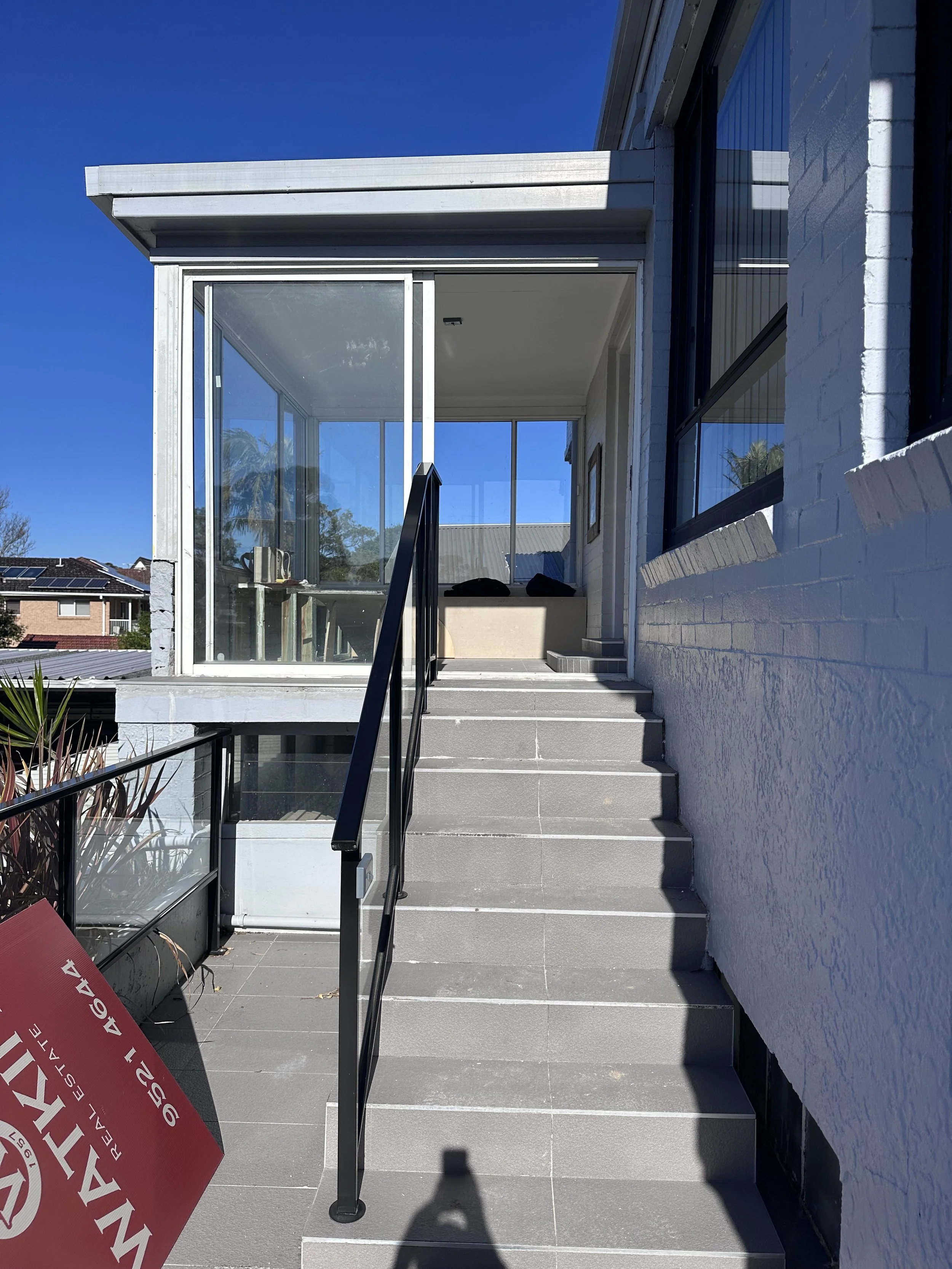 Exterior view of a modern house with stairs leading up to a glass-enclosed porch area, white brick wall, black metal railing, and a clear blue sky.
