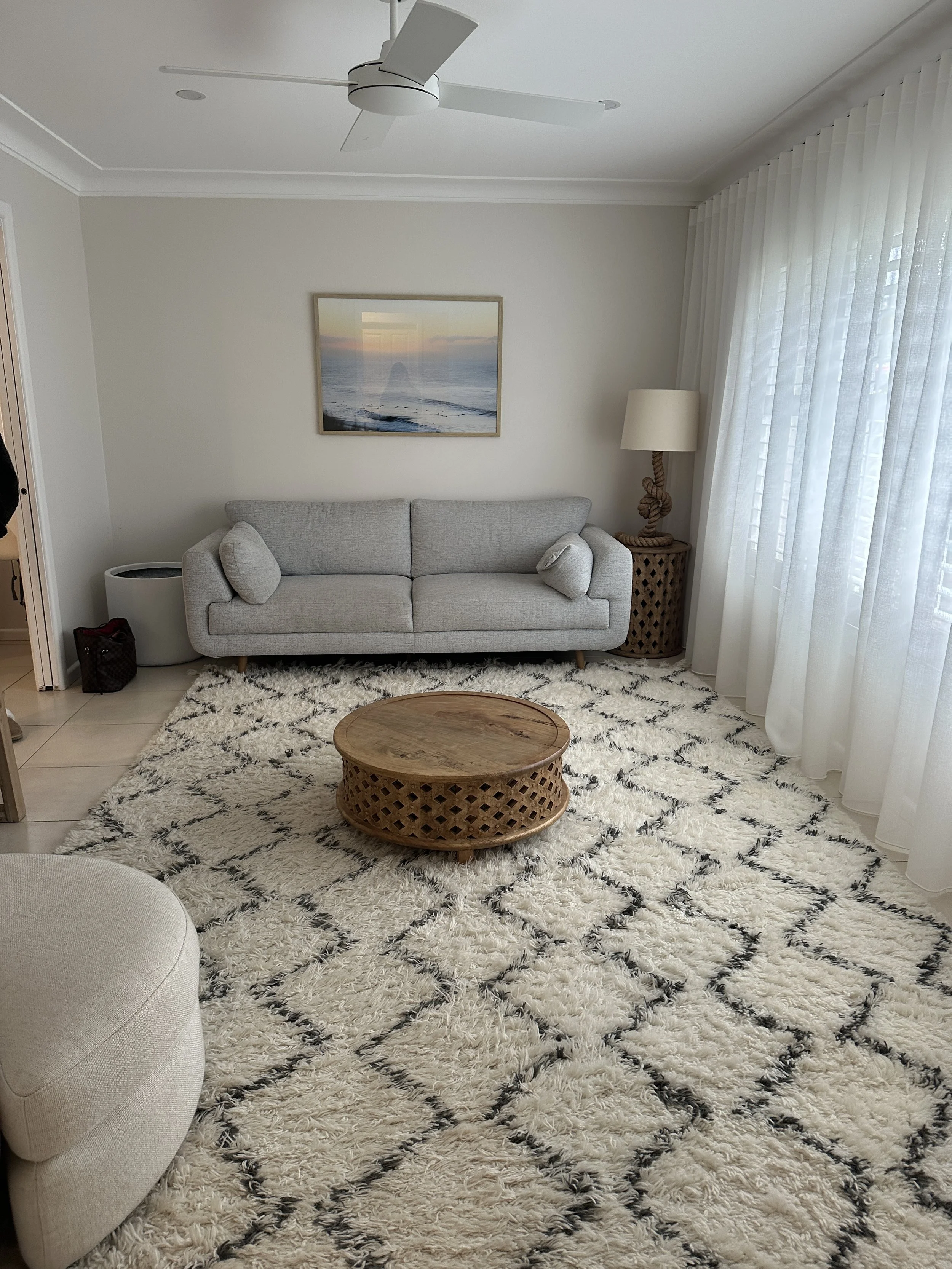 Living room with a gray sofa, a round wooden coffee table, a black and white patterned rug, and a large window with white curtains. There's a floor lamp with a beige shade and a picture of a sunset over the ocean on the wall.