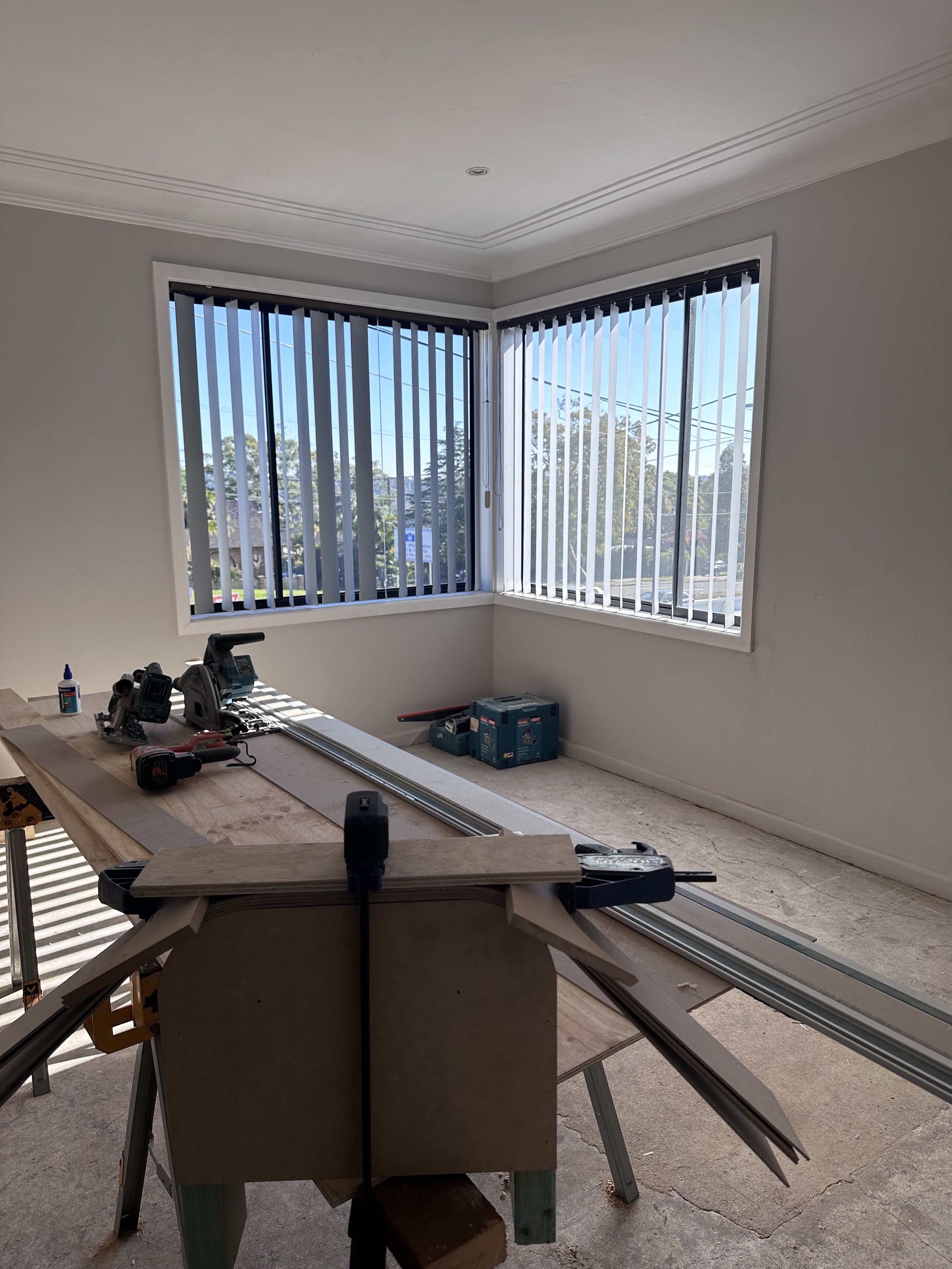 Interior room under renovation with a large window covered with vertical blinds, construction tools on a workbench, and materials for building or carpentry.