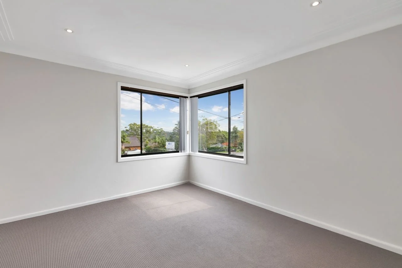 Empty room with white walls, gray carpet, and two large windows showing a suburban neighborhood with trees and houses outside.