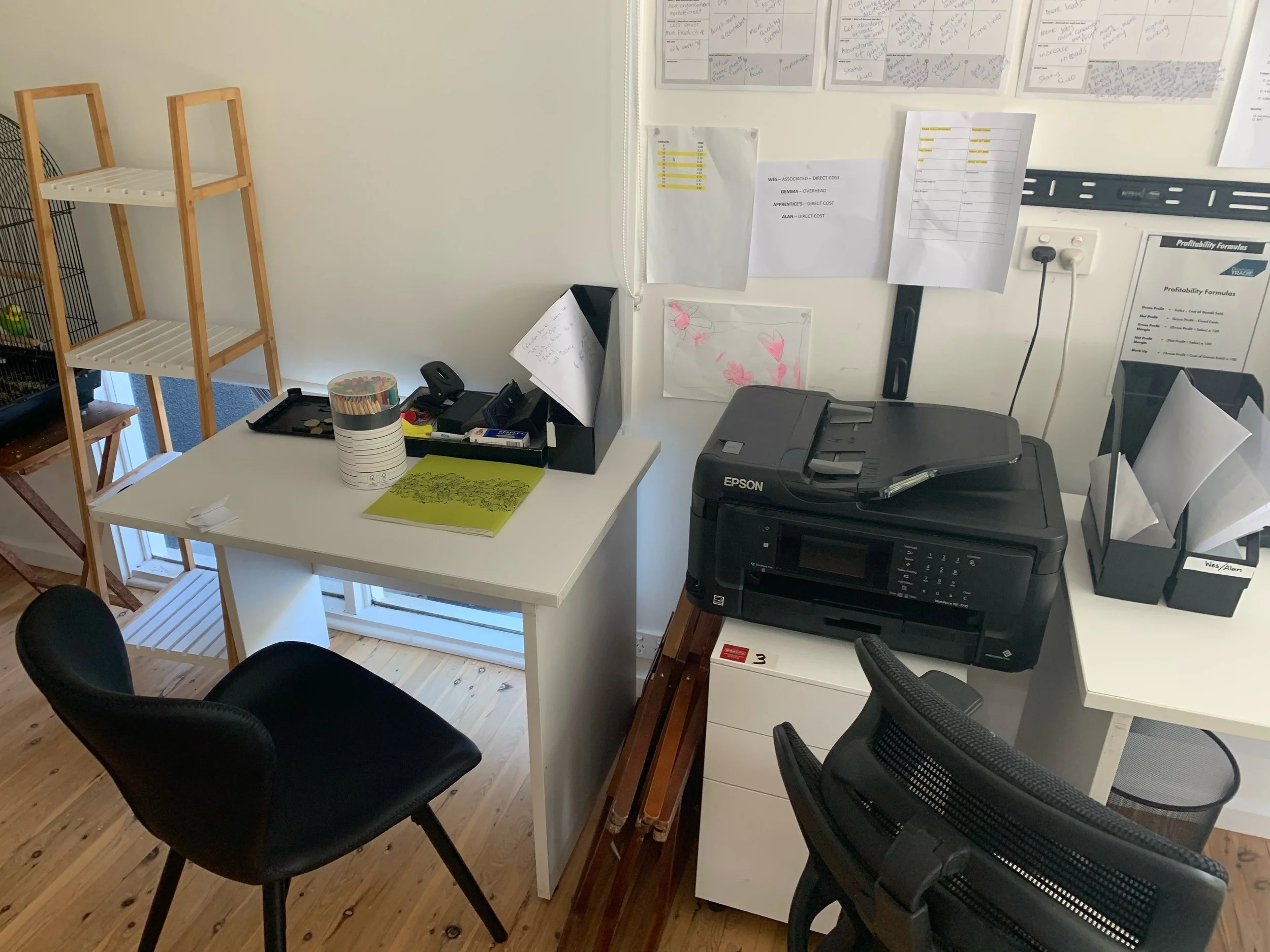 Office workspace with white desks, black office chairs, a printer, a stack of papers, and a small wooden shelf. The wall is decorated with papers, charts, and notes.