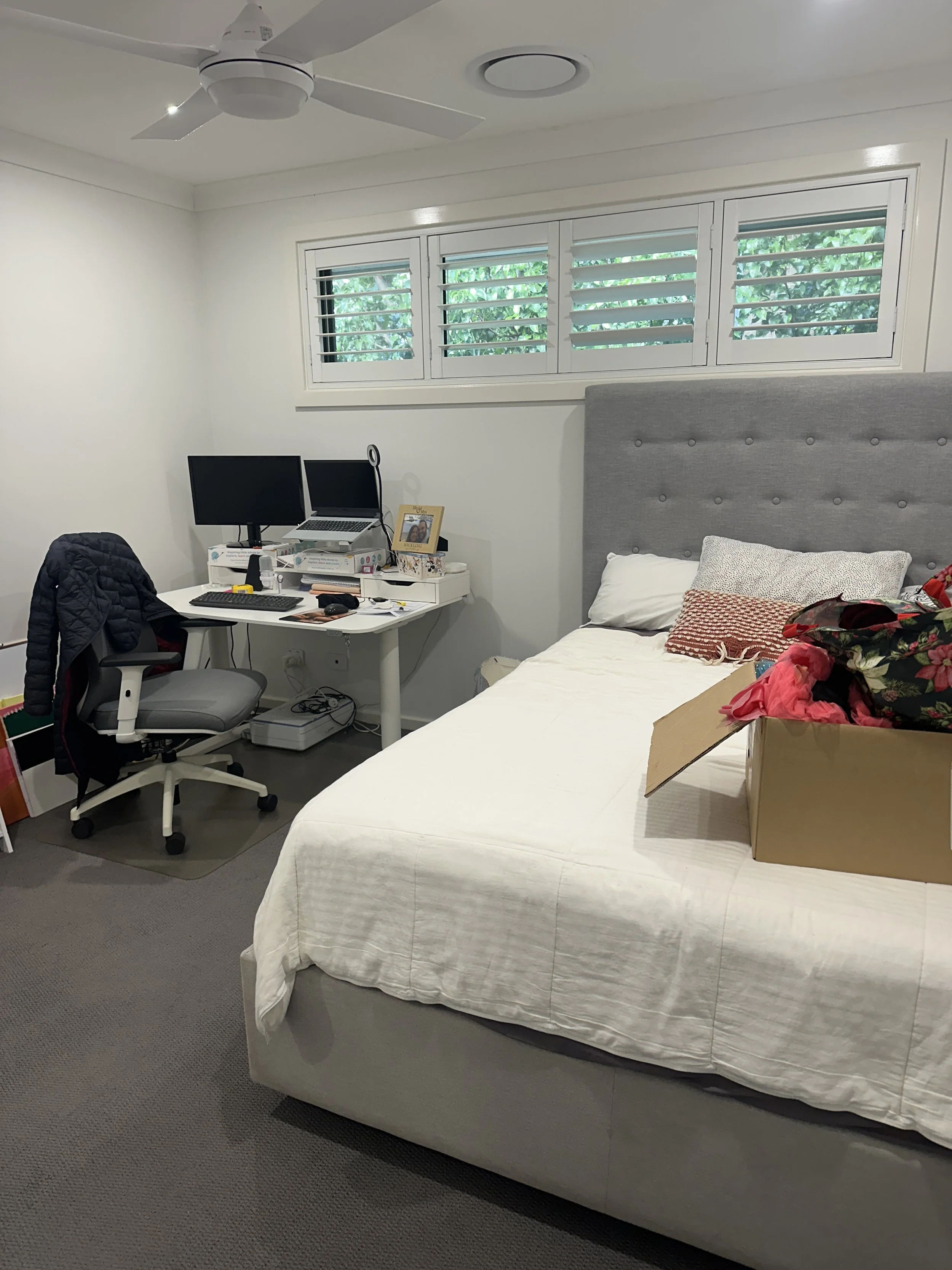 A bedroom with a bed, a dresser, a window with shutters, and a small workspace with two monitors, a laptop, and various items on the desk.