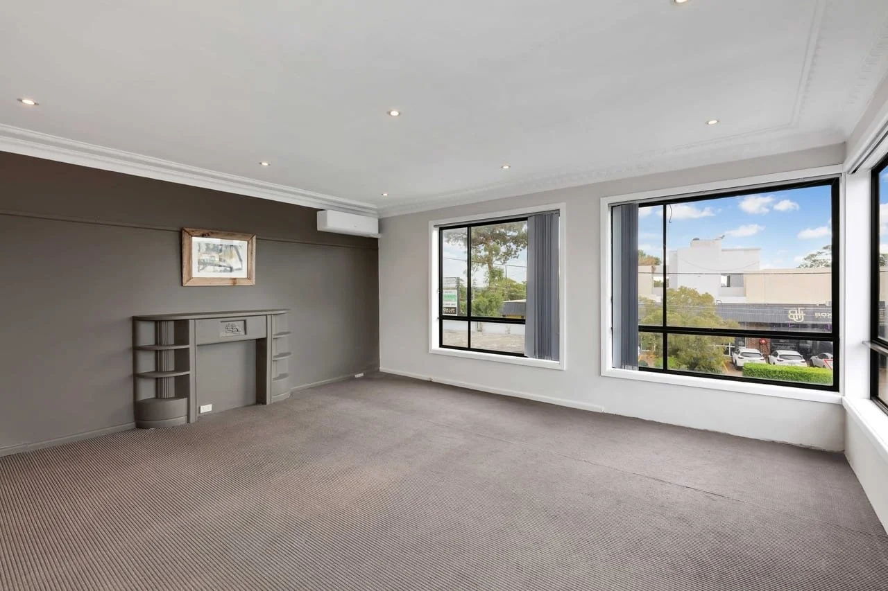 Empty living room with large windows, beige carpet, gray accent wall, and fireplace with shelves, during daytime.