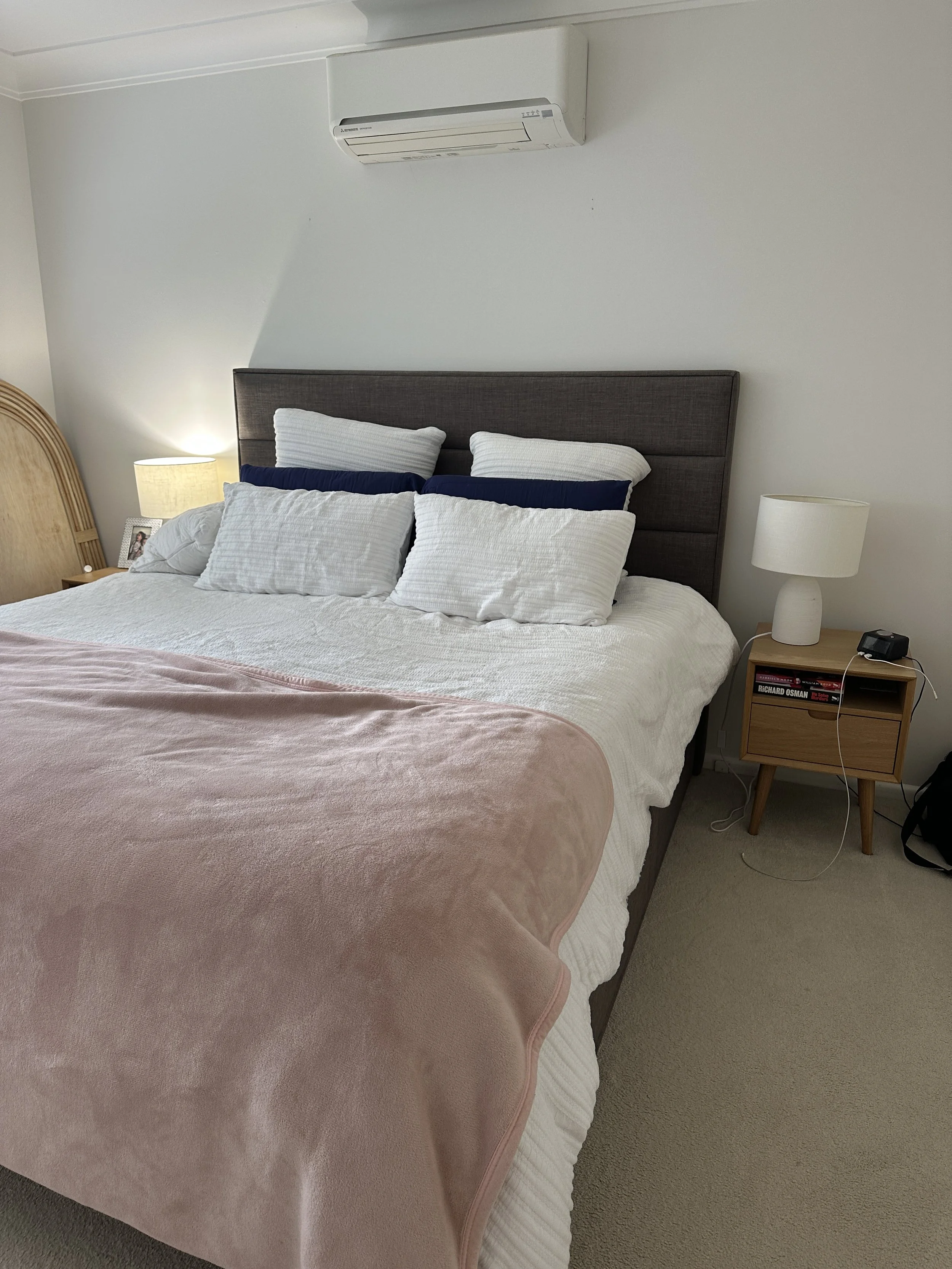 A bed with white bedding, several pillows, and a pink blanket. There are two bedside tables, each with a lamp. An air conditioning unit is mounted on the wall above the bed.