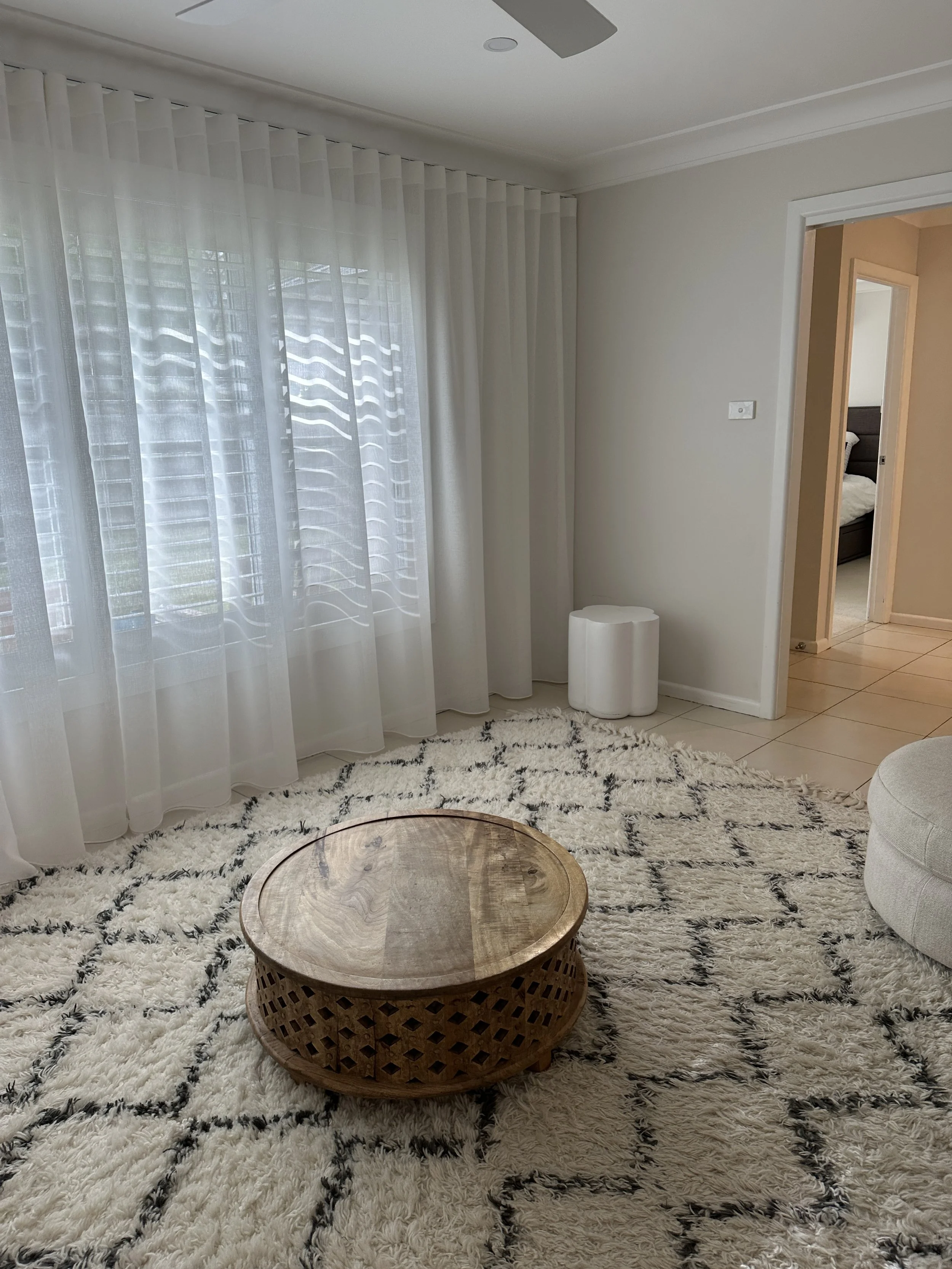 Living room with white sheer curtains covering a large window, a white shaggy rug, a round wooden coffee table, and an off-white sofa armrest visible on the right side. There is a doorway to a bedroom in the background.