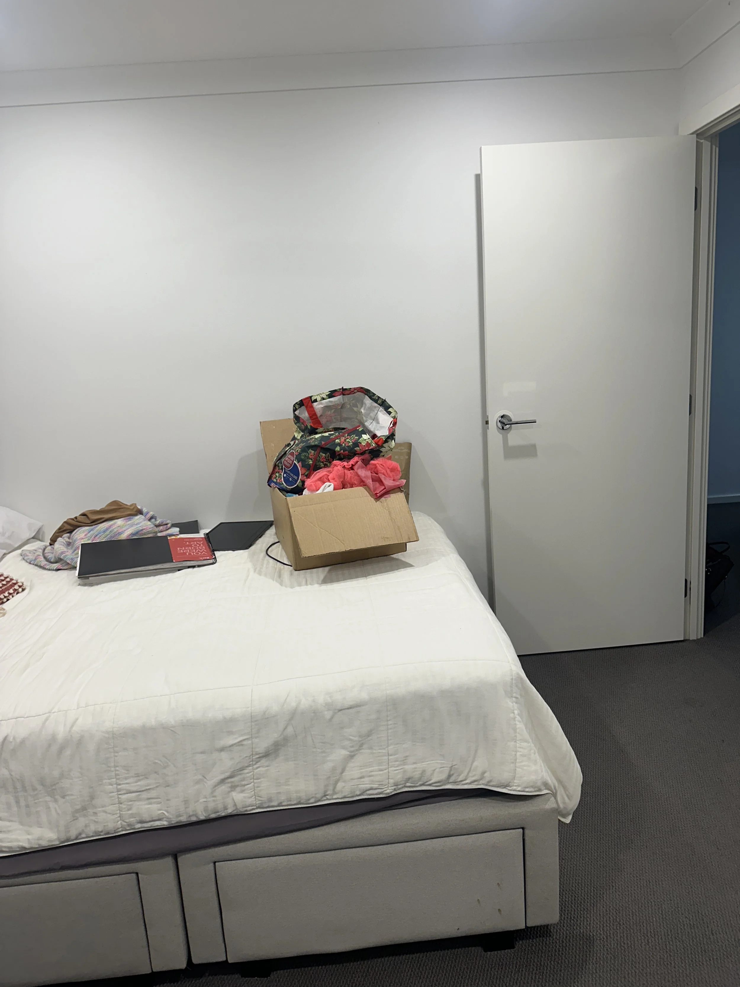 A hotel room with a white bed, some books, clothes, and electronics on it, a cardboard box with clothing and a bag on top, a white door slightly open, and a blue wall visible through the door opening.