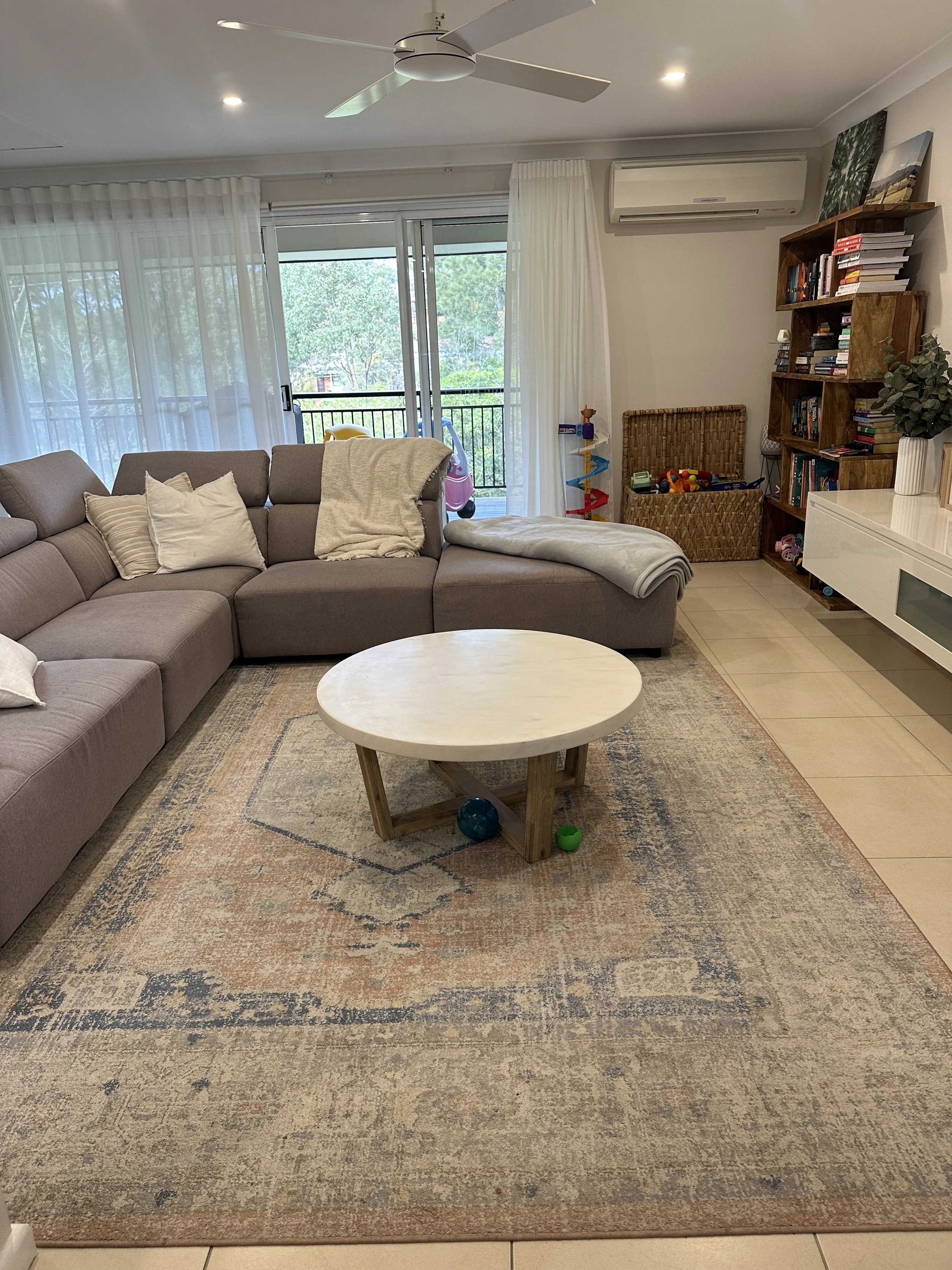 Living room with beige sectional sofa, white pillows, and blanket, wooden coffee table on a patterned rug, sliding glass door with white curtains opening to a balcony with trees, bookshelf with books and toys, white TV console, potted plant, and air 