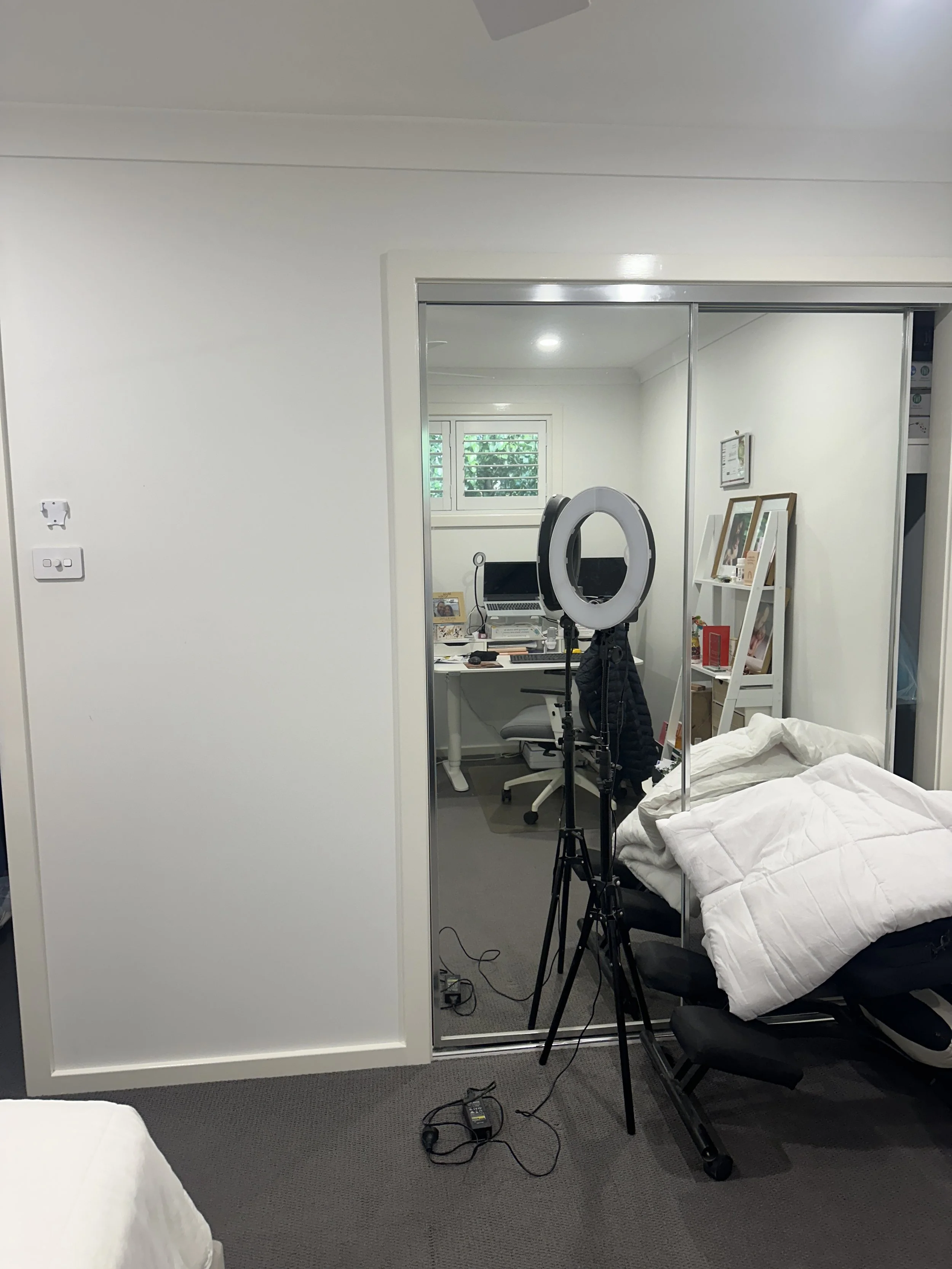 A bedroom with a bed, chairs, a mirror, and a ring light set up for filming or photography. There are various items and frames on the shelves and a window with blinds.