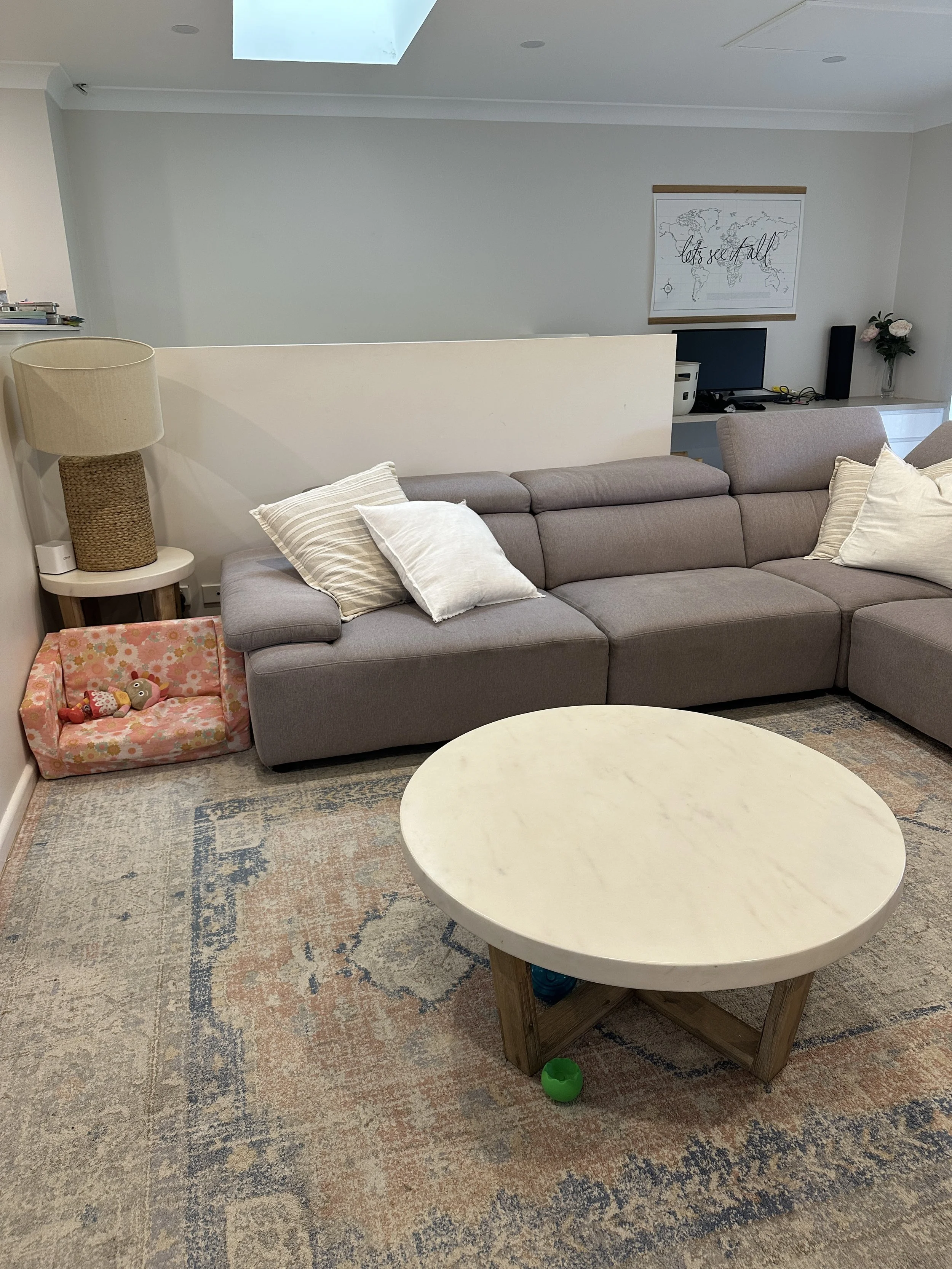 Living room with a gray sectional sofa, pillows, a round white coffee table, a beige lamp, a toddler's bed with stuffed toys, a patterned area rug, a wall decor with a world map, a TV, and a vase with flowers.
