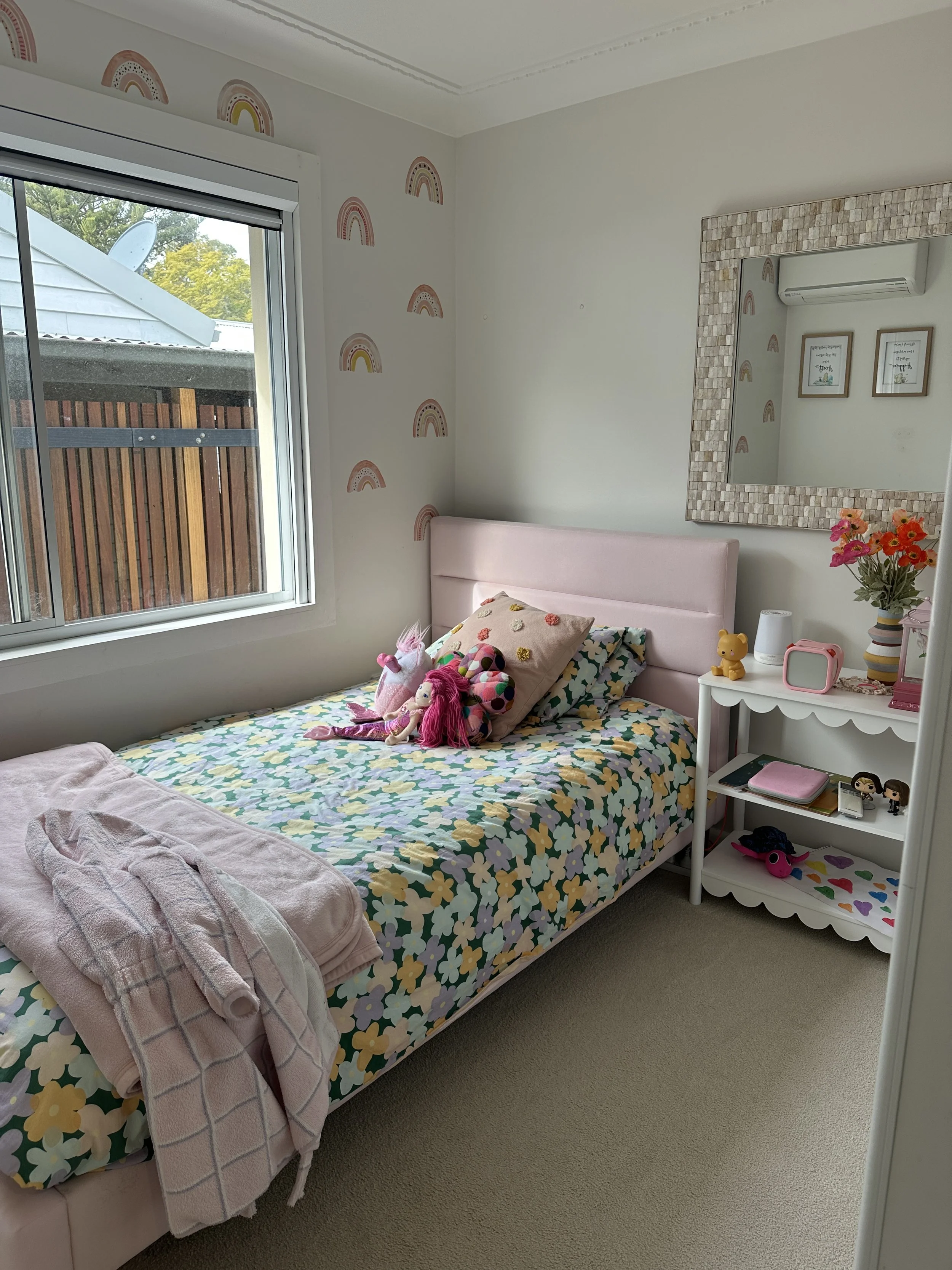 A cozy girl’s bedroom with a single bed covered in a floral quilt, stuffed animals, and pillows. There is a window with a view of a backyard with trees and a fence. The room has pastel-colored decor, including rainbow wall decals, a large mirror with